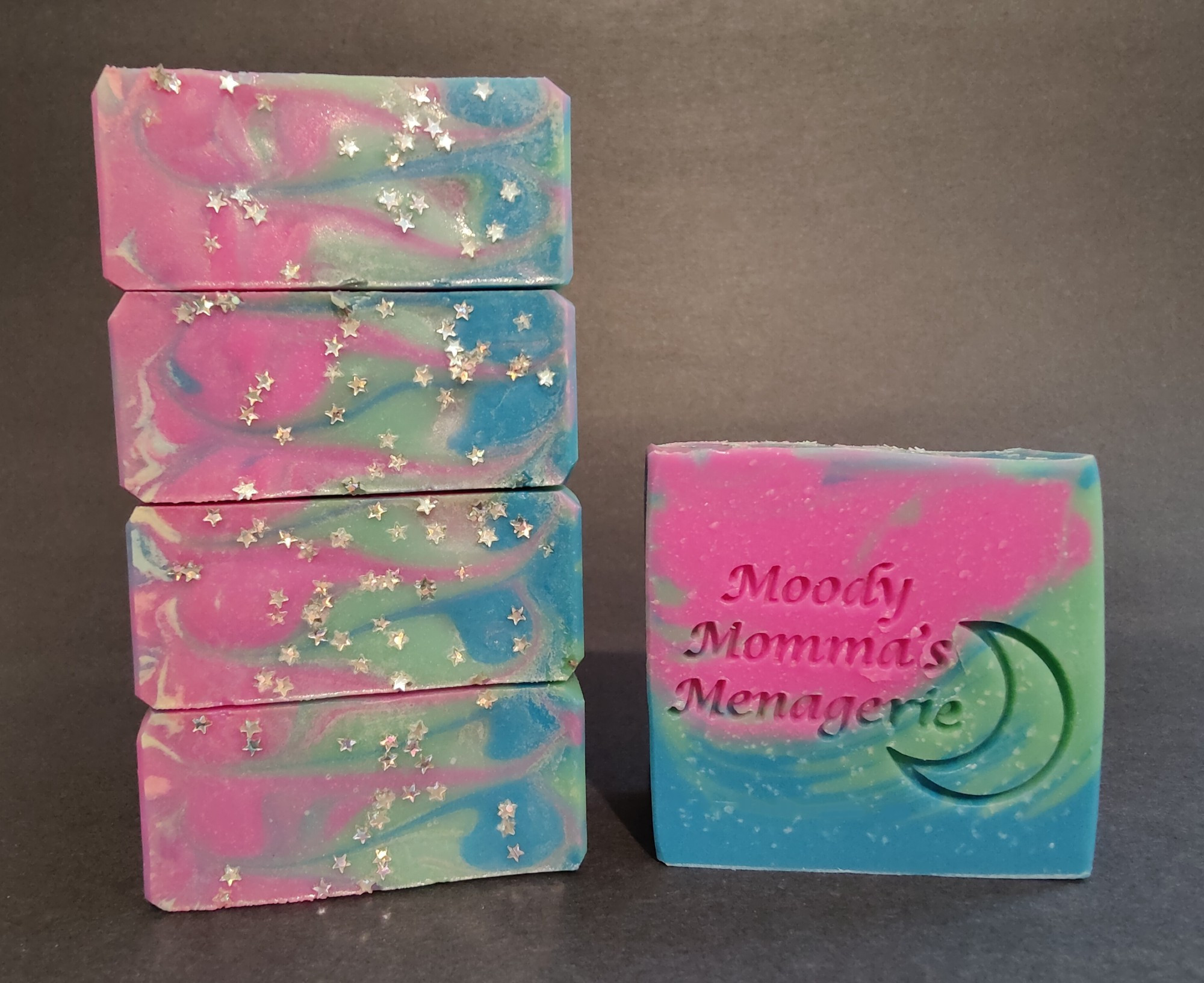 On the left are four bars of pink, blue and green soap stacked on their side to show the top detail with star eco glitter. The right bar is stamped with "Moody Momma's Menagerie" and a crescent moon