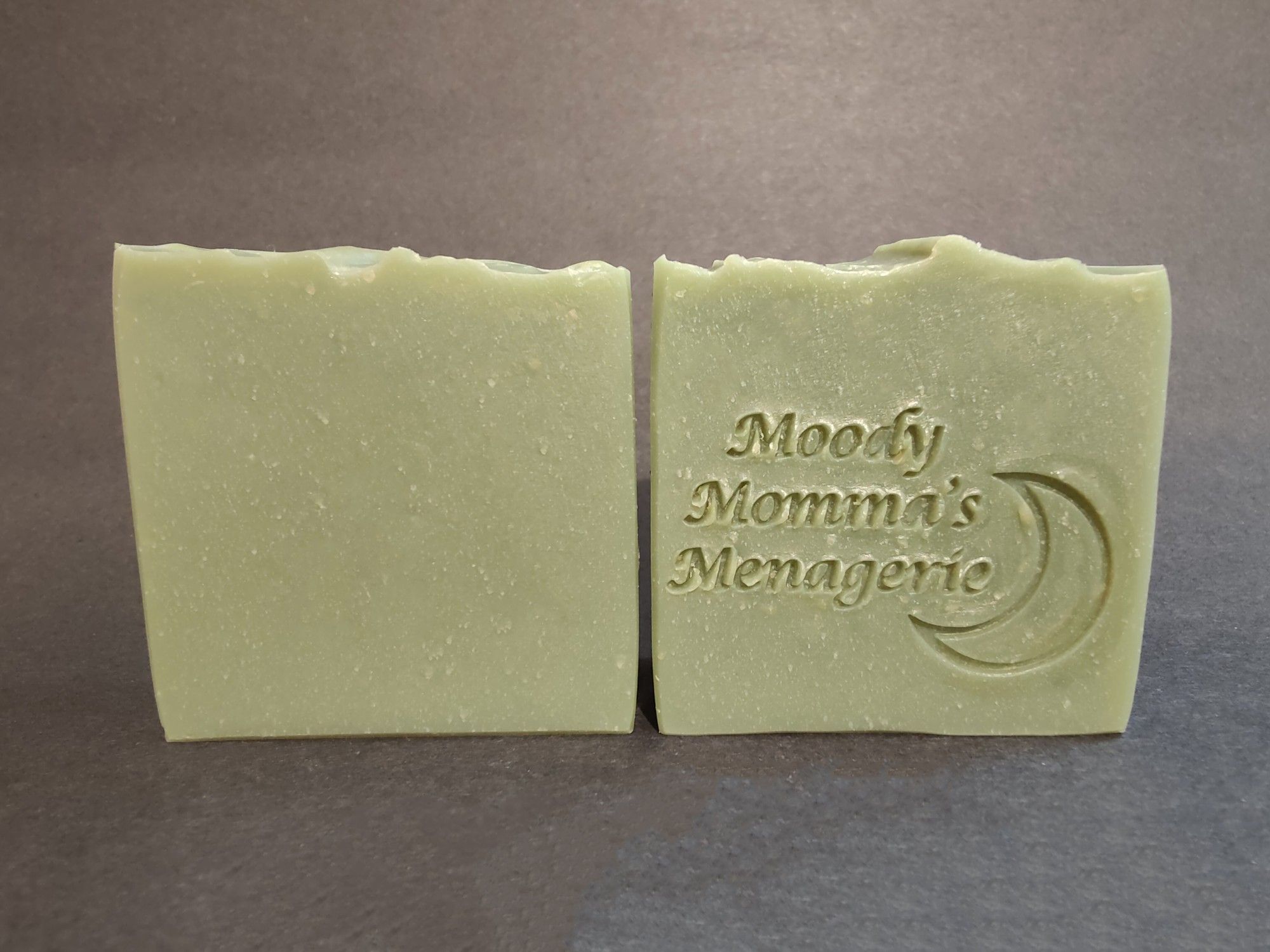 Two bars of green soaps sitting side by side. The right bar is stamped with "Moody Momma's Menagerie" and a crescent moon