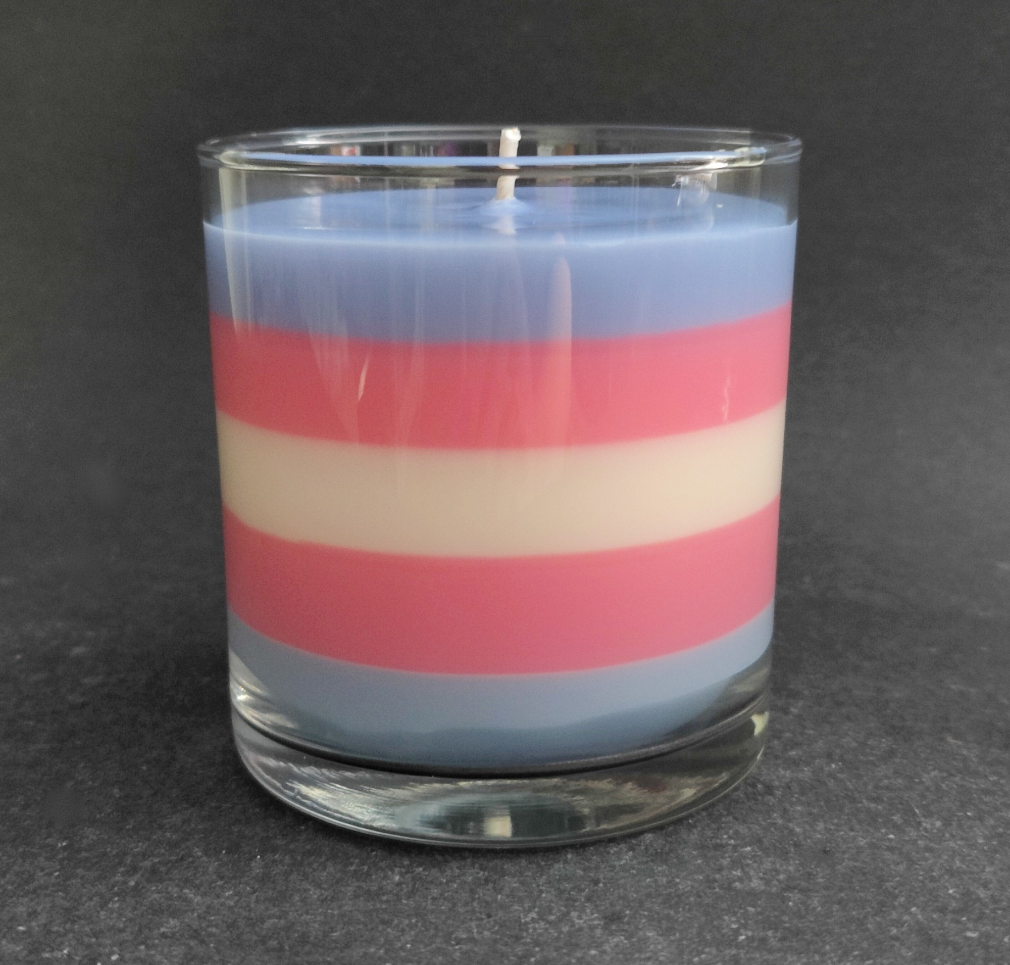 A candle in a glass status jar layered with blue, pink, white, pink and blue to represent the Transgender Pride flag