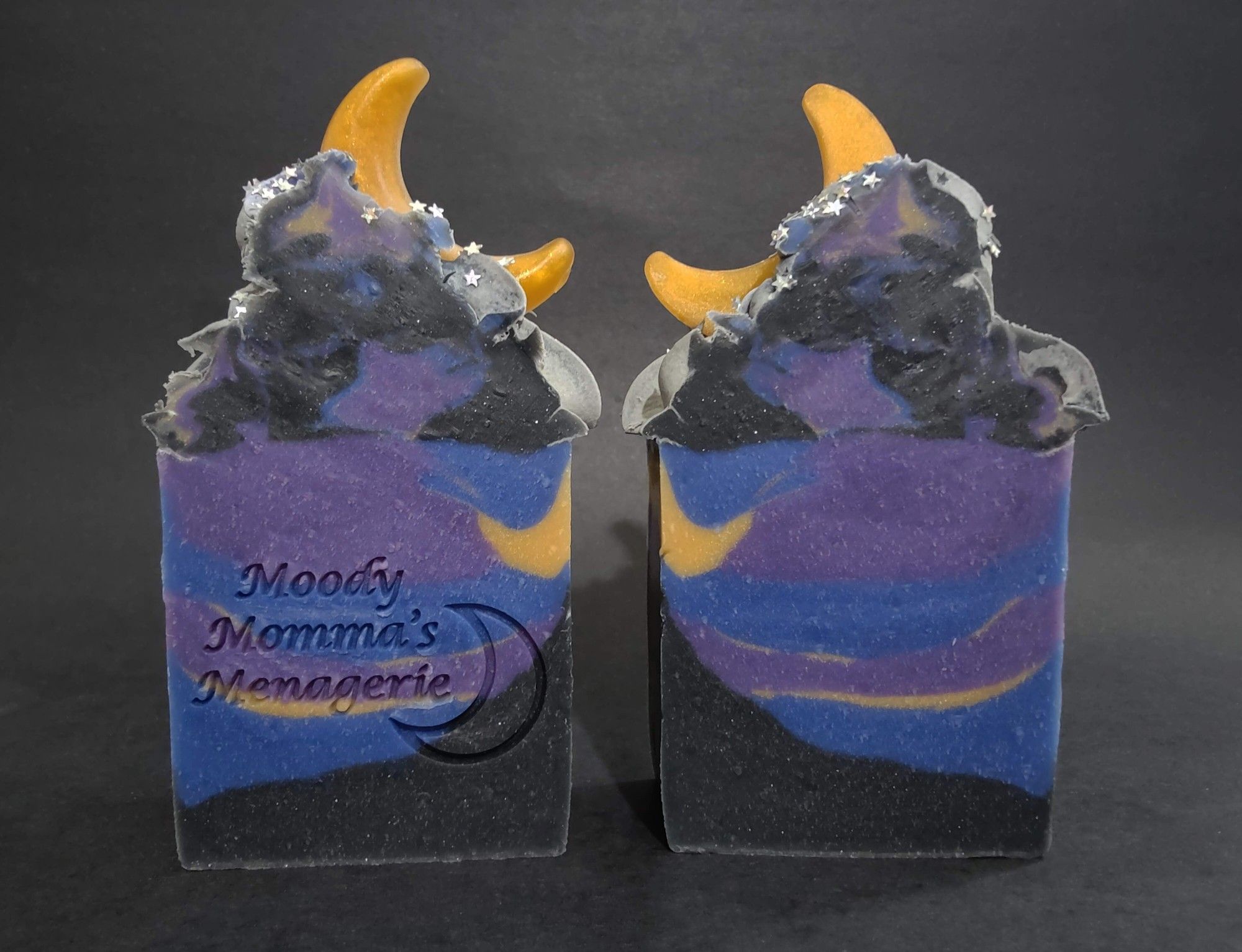 Two bars of black, blue, purple and gold soaps inspired by Starry Night, with a gold crescent moon soap on top and star shaped, plant based, biodegradable glitter. The left bar is stamped with "Moody Momma's Menagerie" and a crescent moon. These soaps are called Shooting Stars and scented a luxurious and complex blend of lavender and cedar leaf, with warm notes of amber, vanilla, sandalwood, lily of the valley and a woodsy sweet rosewood base.