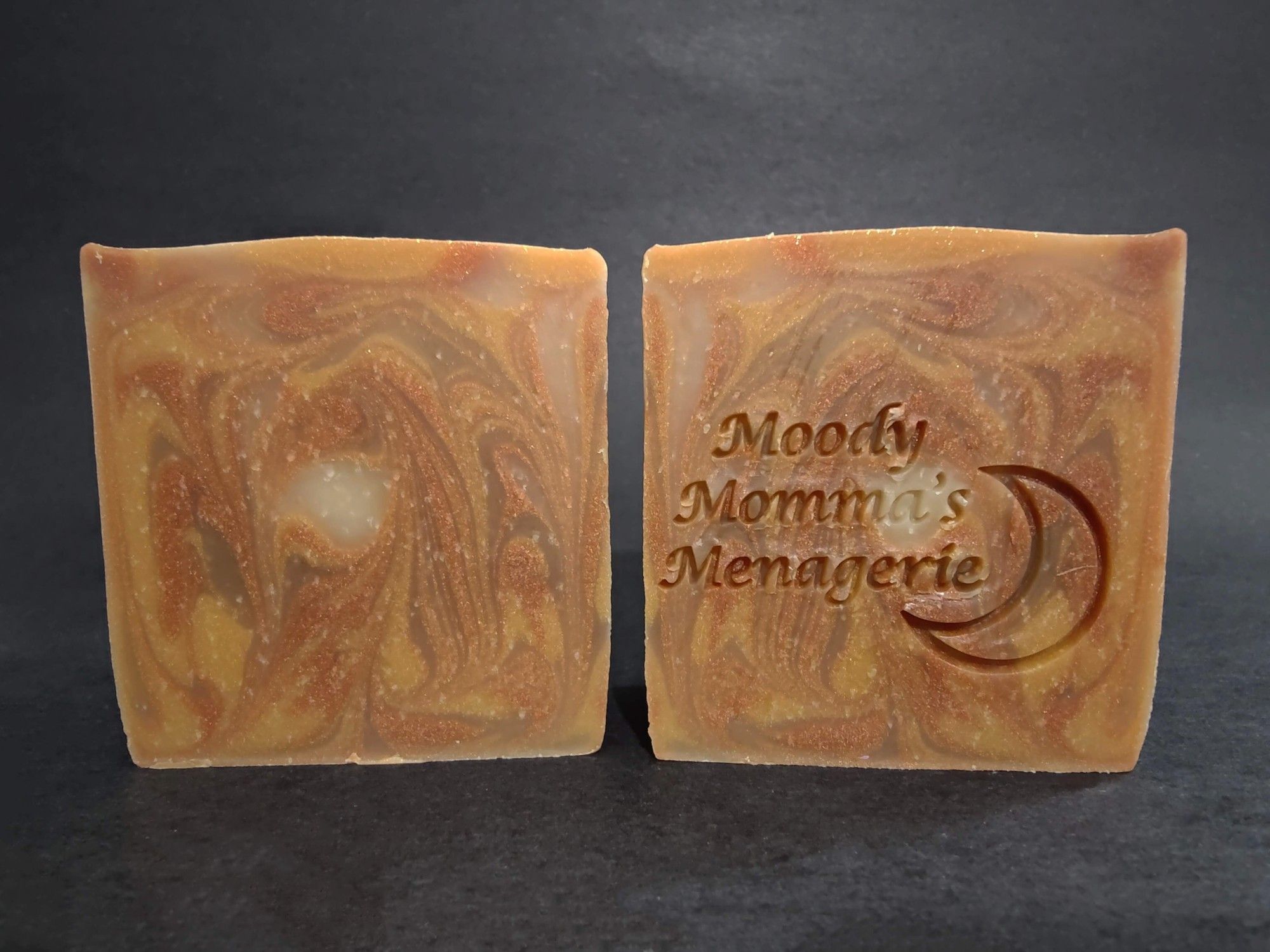 Two bars of bronze, copper and cream swirled soaps sitting next to each other. The bar on the right is stamped with "Moody Momma's Menagerie" and a crescent moon