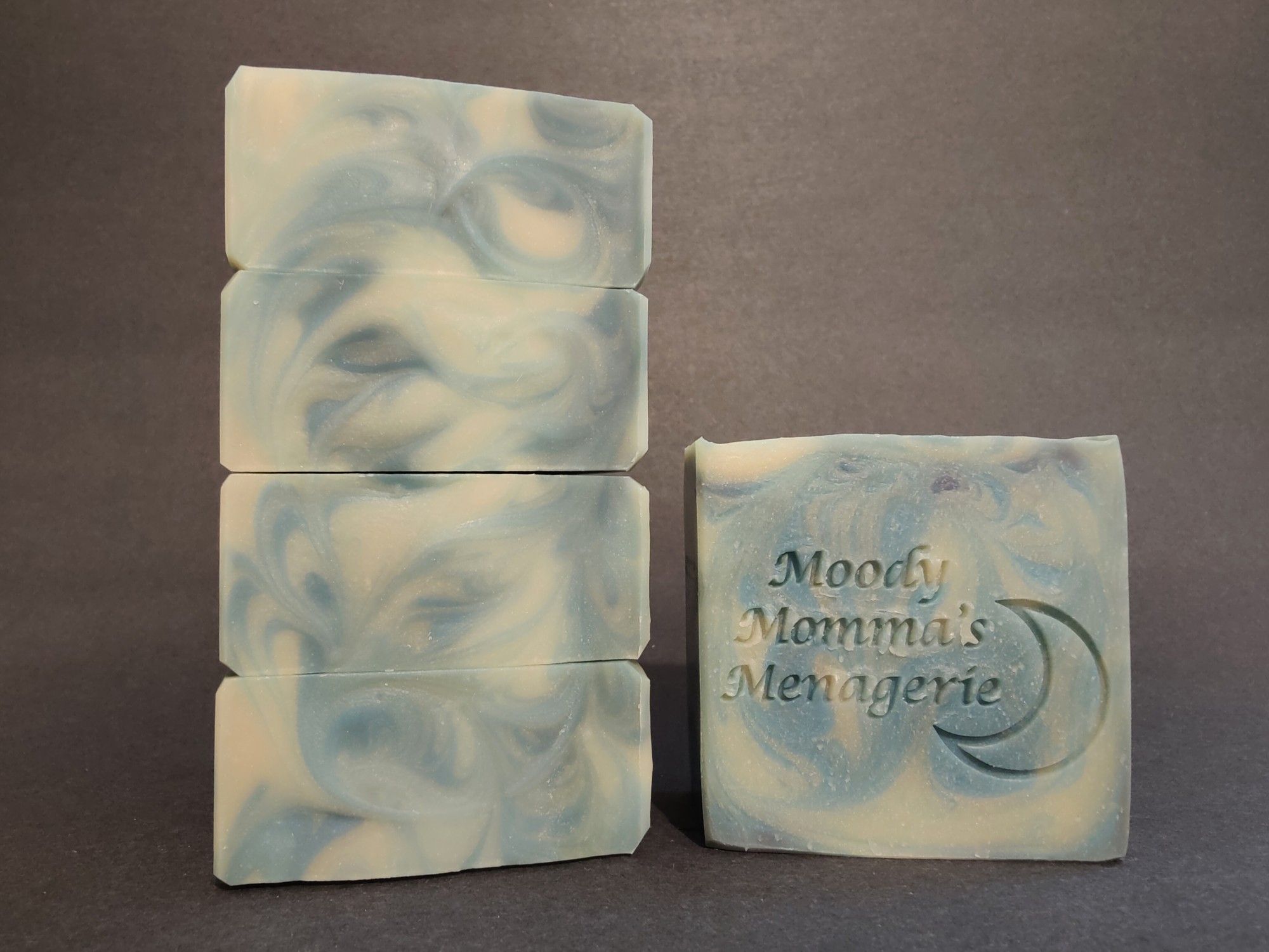 On the left four blue and white swirled soaps are stacked on their side to show the wispy top detail. On the right is a single bar of blue and white swirled soap with a crescent moon and "Moody Momma's Menagerie" stamped into it