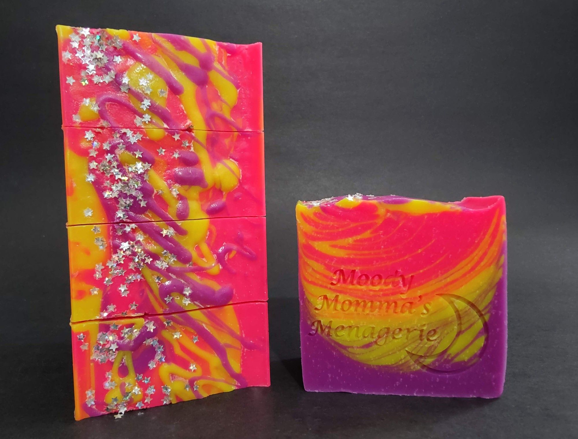 On the left are four bars of neon pink, yellow and purple soaps stacked on their side to show the top paint splatter like design with plant based star shaped glitters. On the right is a single bar of the soap stamped with "Moody Momma's Menagerie" and a crescent moon