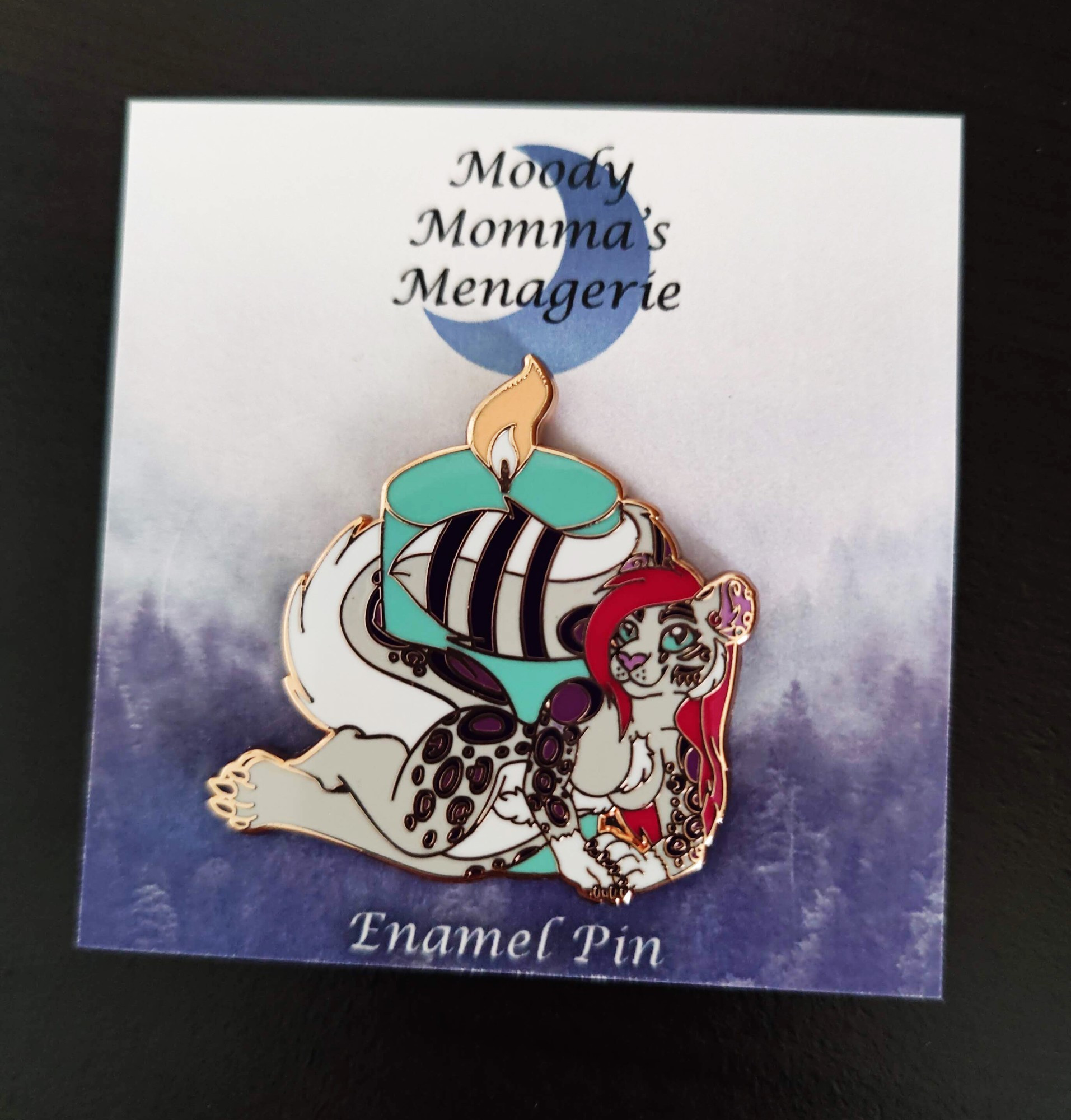 An enamel pin of my fursona, a white and grey snow leopard with purple spots, raspberry colored hair and blue eyes, laying in front of a blue candle. The card has my moon logo with "Moody Momma's Menagerie" and "enamel pin" printed at the bottom