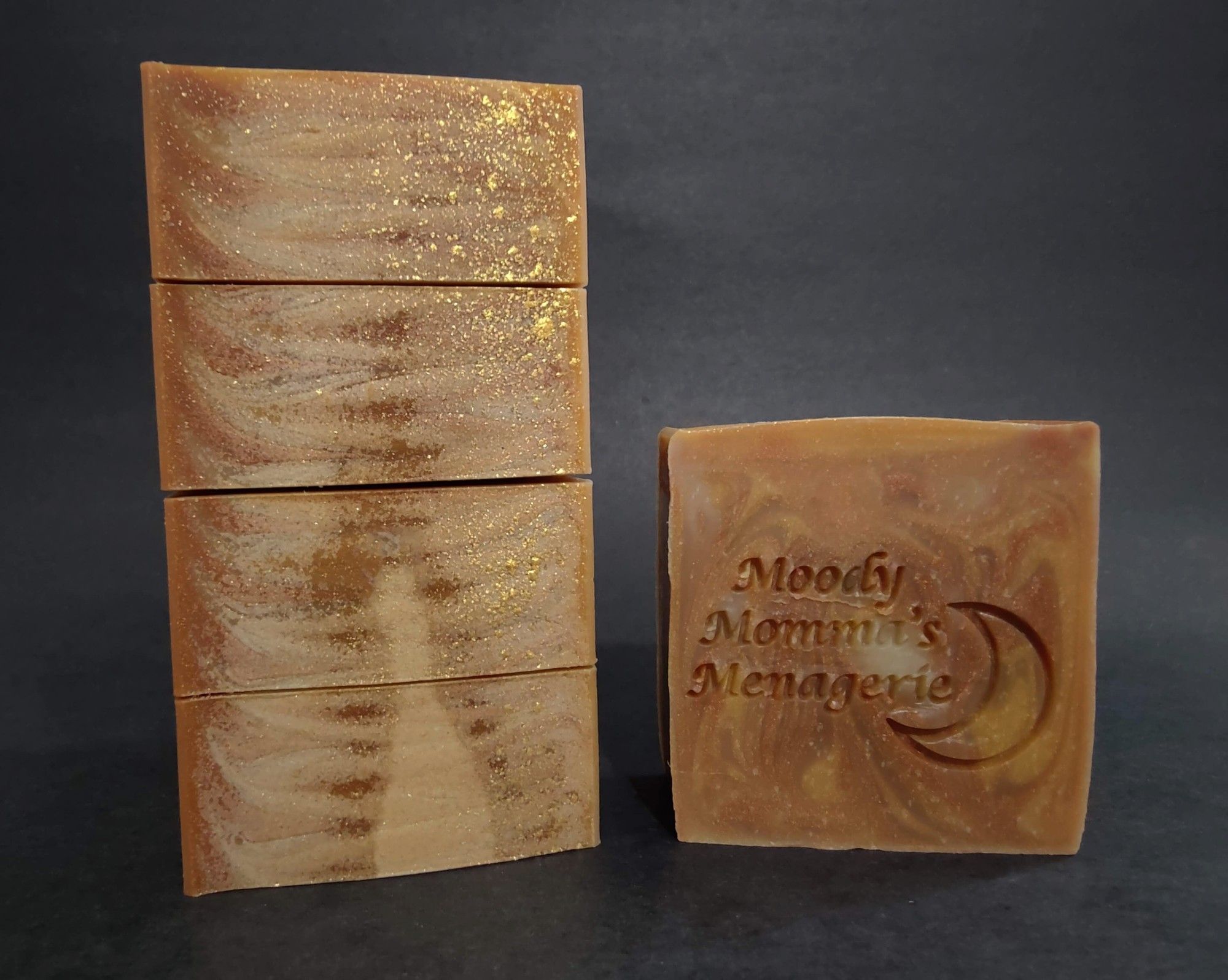 On the left four bars of brown, copper and cream soap are stacked on their side to show the gold sparkle mica on top and a faint swirl. On the right is a single bar of soap stamped with "Moody Momma's Menagerie" and a crescent moon