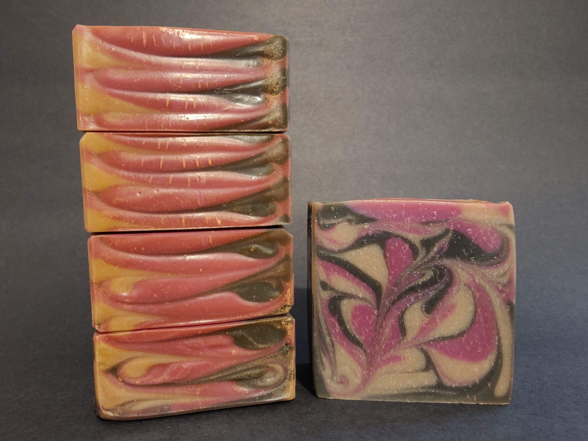 On the left are four bars of cream, black and raspberry colored soaps stacked on their side to show the top detail. On the right is a single bar of the soap to show the swirl pattern