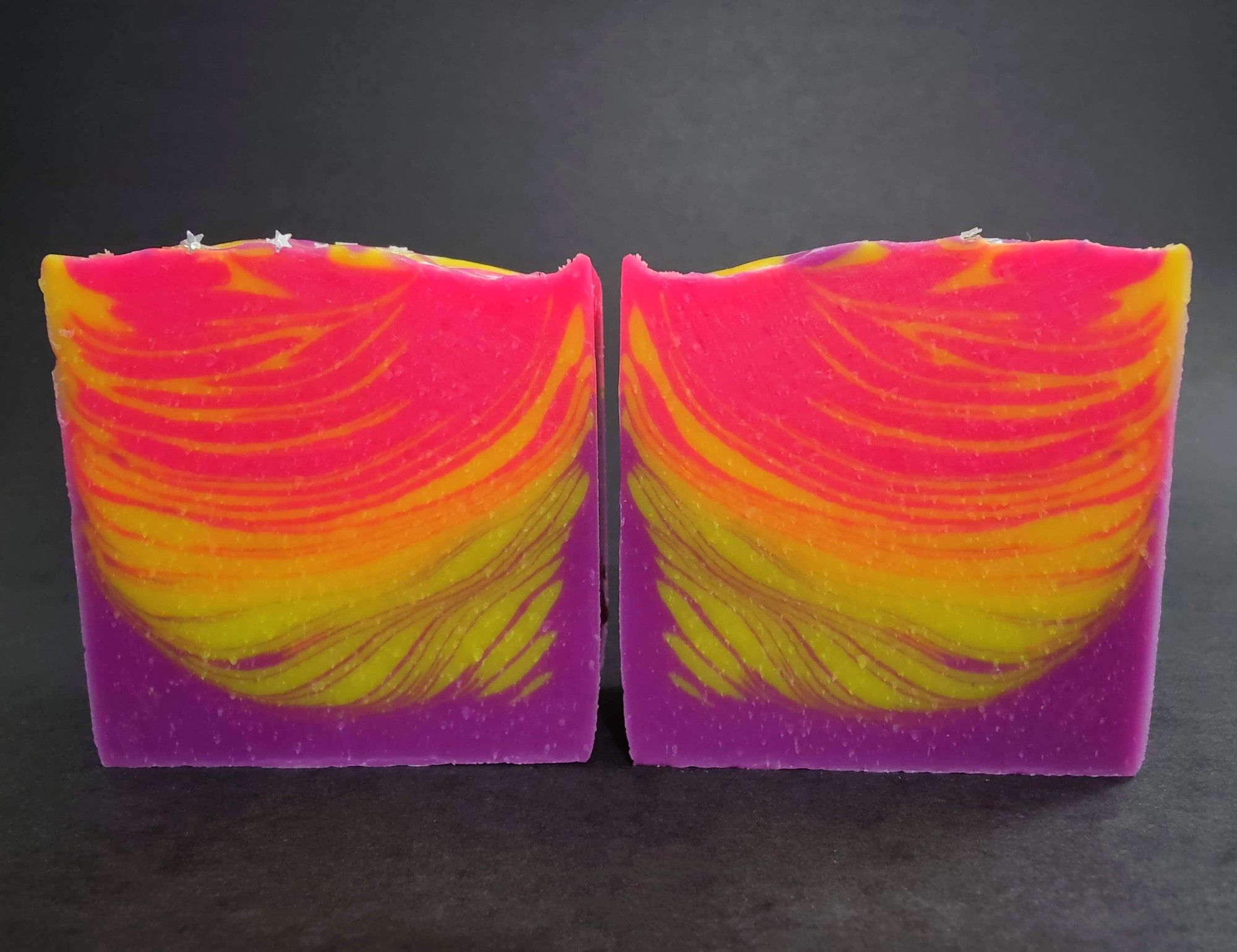 A neon pink, yellow and purple soap with a feathered design and star shaped, plant based, biodegradable eco glitter peeking out from on top. This soap is called Atomic Sunrise and scented with a magical blend of Japanese grapefruit and mandarin, peaches, plums, Valencia grapes, and soft rose petals, all on a warm base of Nubian musk.