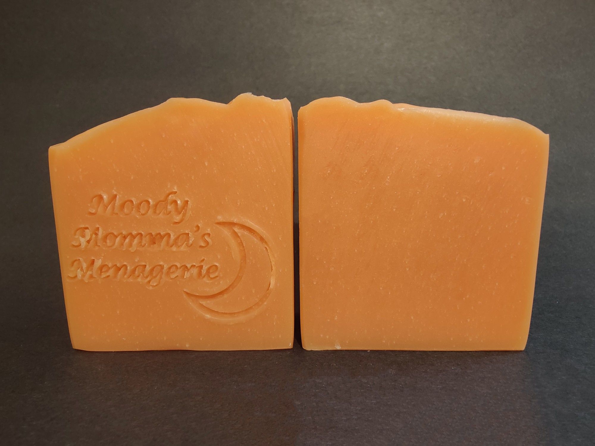 Two bars of orange colored soap sitting side by side. The bar on the left has "Moody Momma's Menagerie" and a crescent moon stamped into it