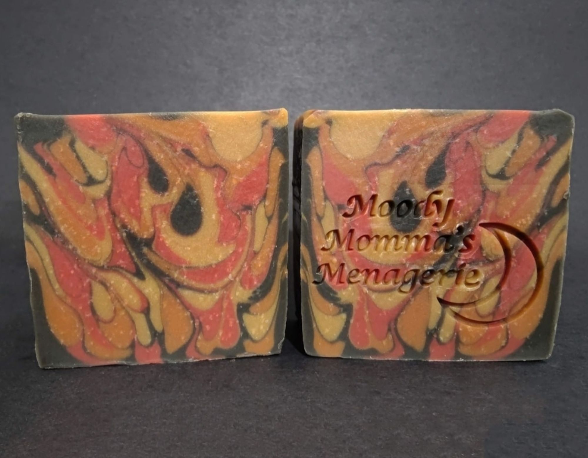 Two bars of red, yellow, orange and black soaps made to look like fire. The right has a crescent moon and "Moody Momma's Menagerie" stamped into it