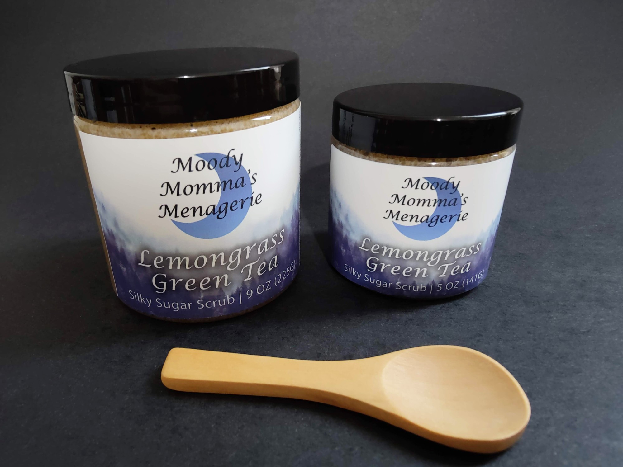 A 9 ounce jar and a 5 ounce jar of sugar body scrubs in Lemongrass Green Tea with a small bamboo spoon in front