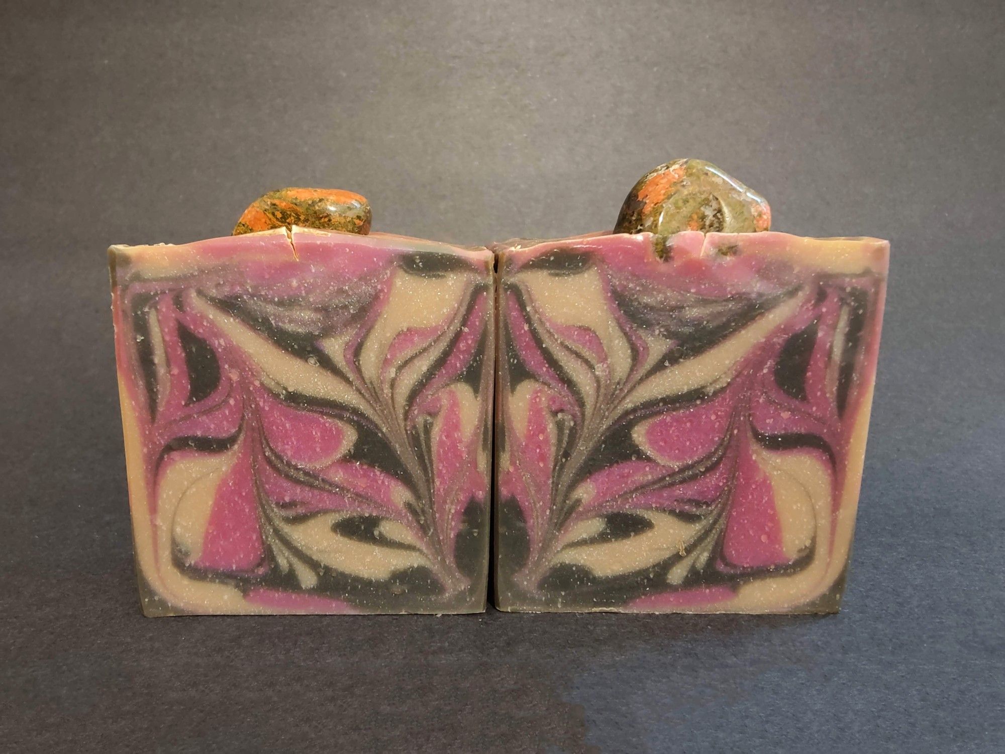 Two bars of cream, black and raspberry colored soaps sitting side by side with a Unakite gemstone tumble on top of each