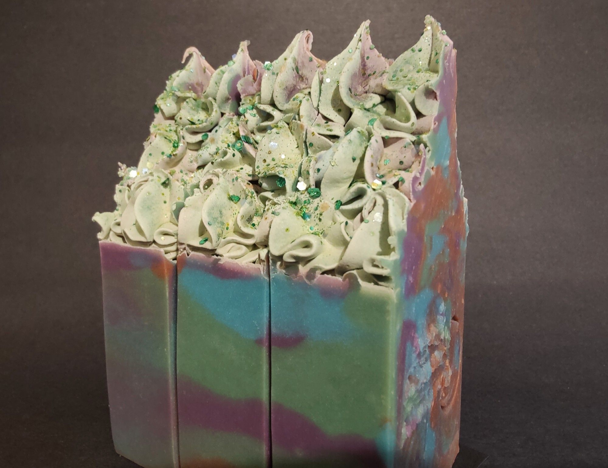 Three large bars of purple, green, blue and brown colored soaps with "frosted" tops and biodegradable plant based glitter, turned to show the frosted details and glitter