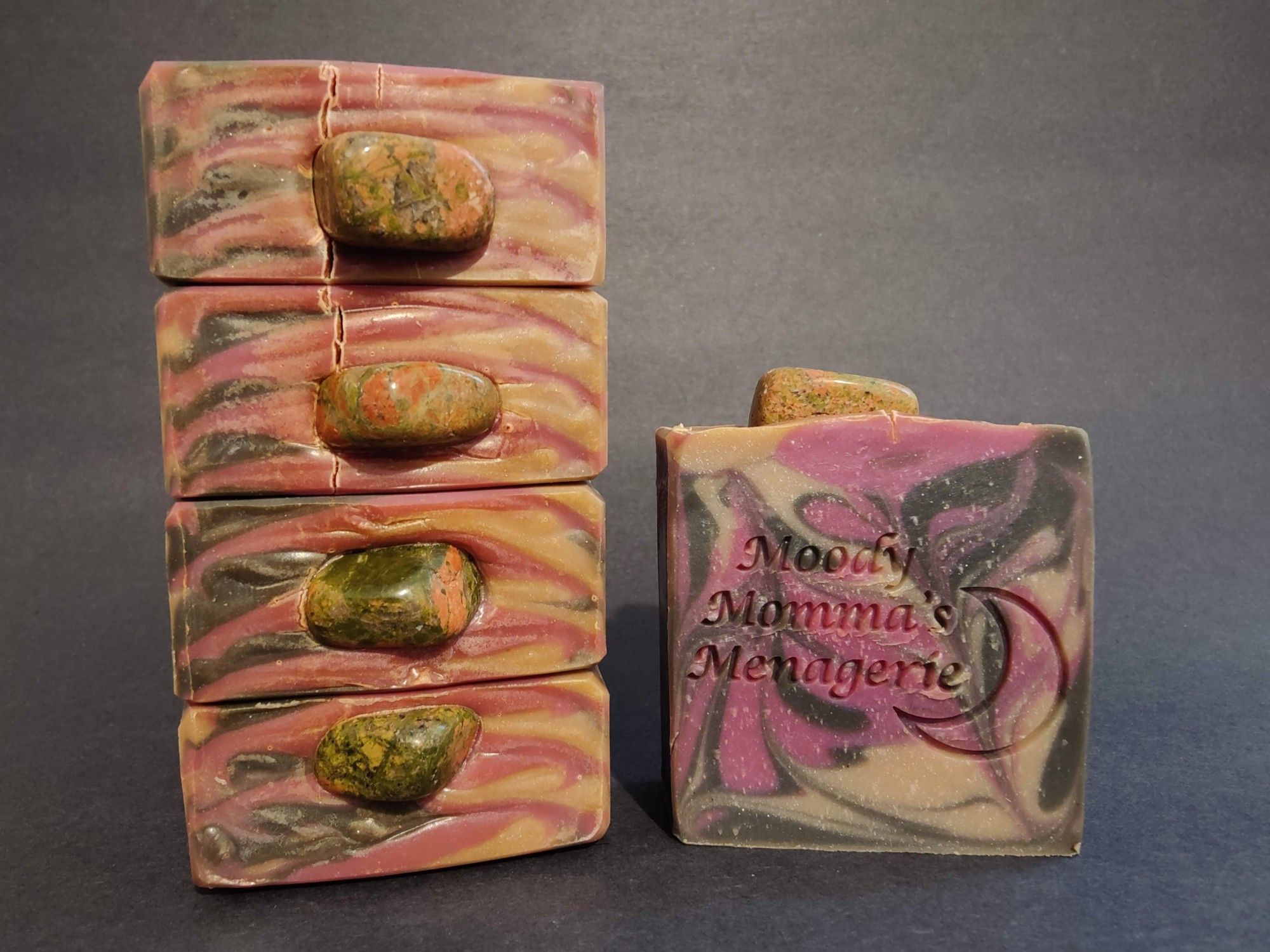 On the left are four bars of cream, black and raspberry colored soaps stacked on their side to show the top detail with a Unakite gemstone tumble placed on each bar. On the right is a single bar of the soap stamped with "Moody Momma's Menagerie" and a crescent moon