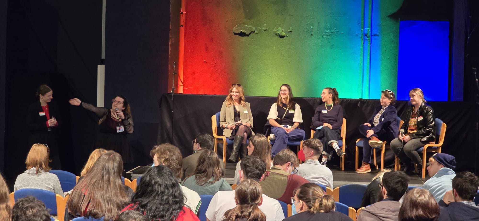 Photo is of the crowd and panel in a dark room with a colourful backdrop