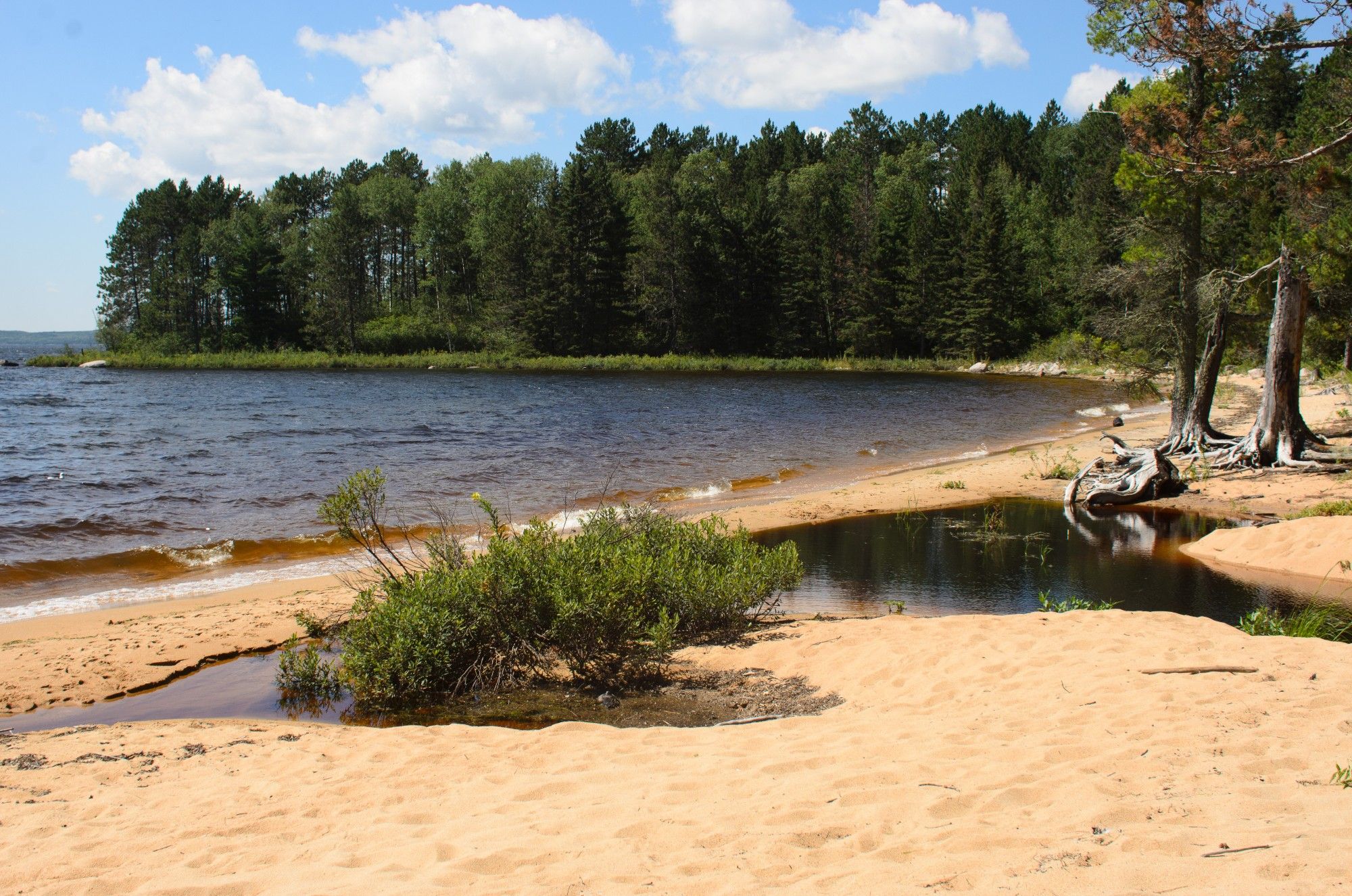 A reddish sand beach in northern Ontario. A creek from the forest almost meets the lake. The forest is spruce, aspen, birch, and pine. The water in the lake is dark with consistent waves. The sky has scattered clouds.