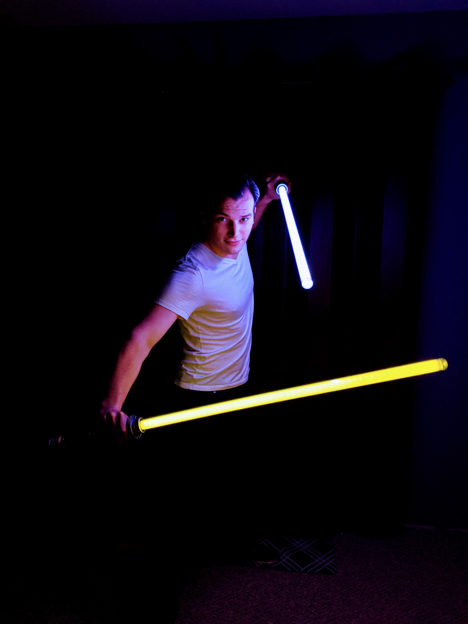 a photo of Colton dual-wielding yellow and white lightsabers. The image is very high-contrast and only his arms, face, and white t-shirt are illuminated by the light of the blades with the rest of the room covered by an inky shadow