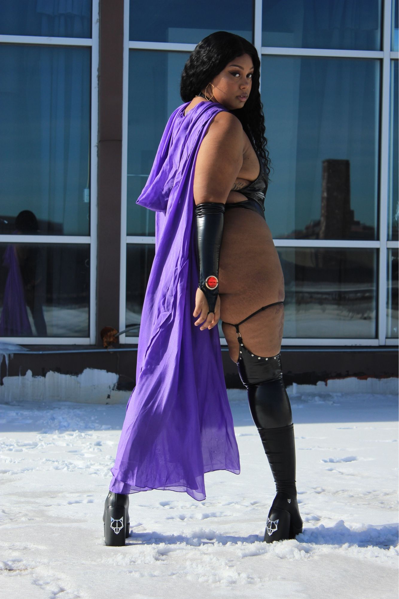 Standing in the snow in platform Naked Wolfe black vegan leather boots. My purple cape is covering the left side of my body. I am standing body facing away from the camera but looking back at it