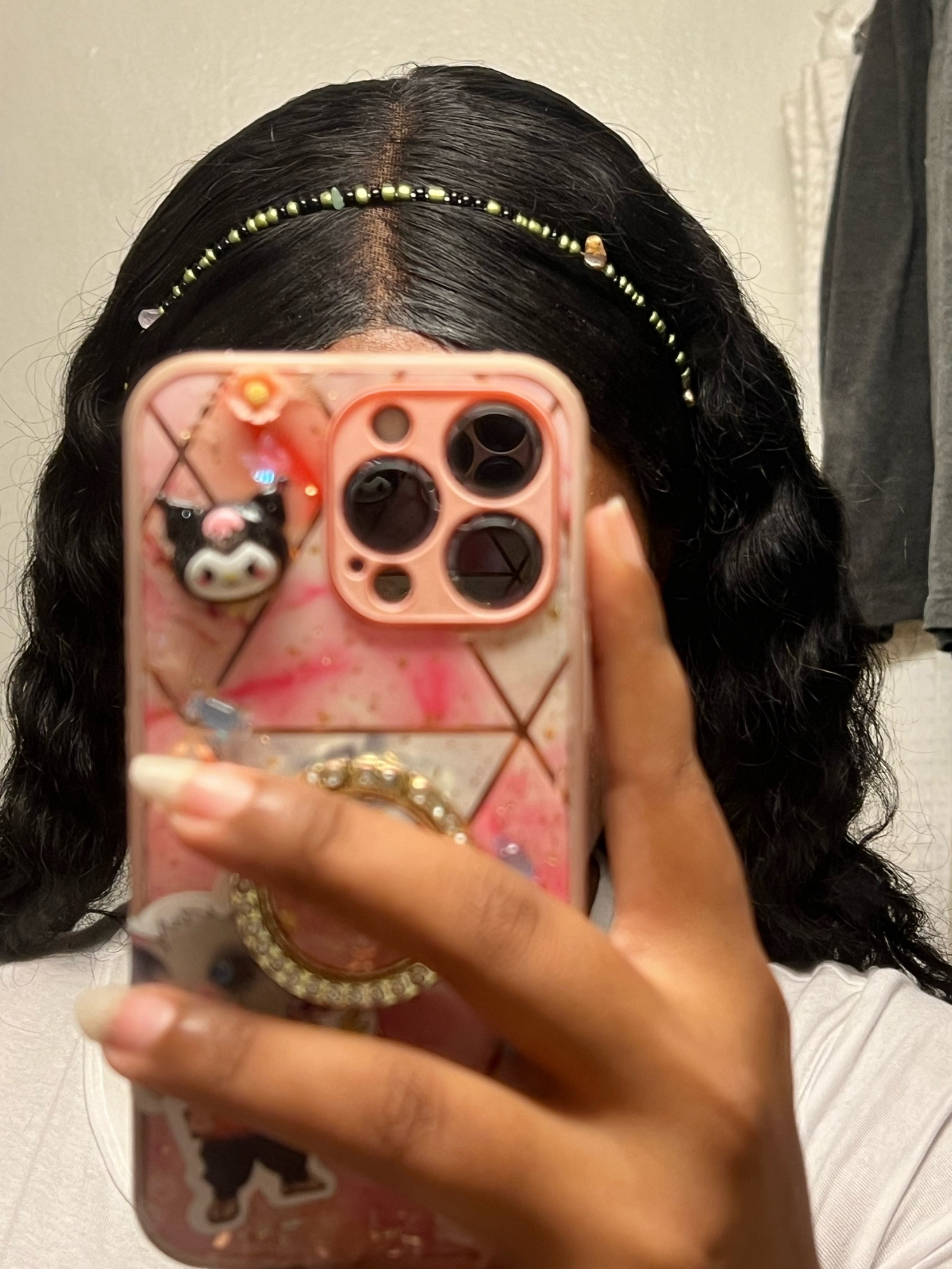 Princess in the mirror but her phone is blocking her face, all that can be seen is a pretty beaded hair band and her black wavy hair. Her phone case is pink with geometrical lines on it as well as a kuromi charm and an inosuke sticker partially covered by her hand