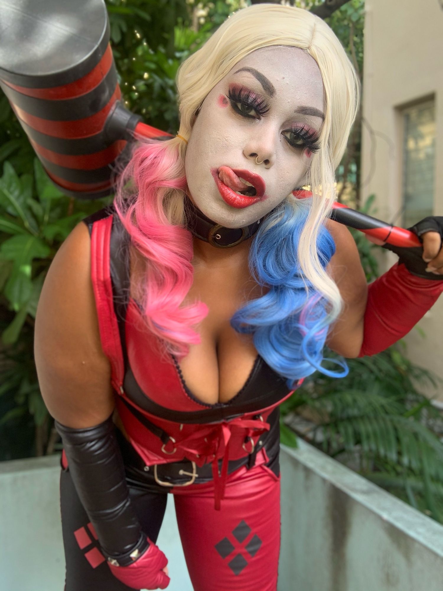 Princess dressed as Harley Quinn Arkham, leaning towards the camera with a hammer their back and licking the side of their lips