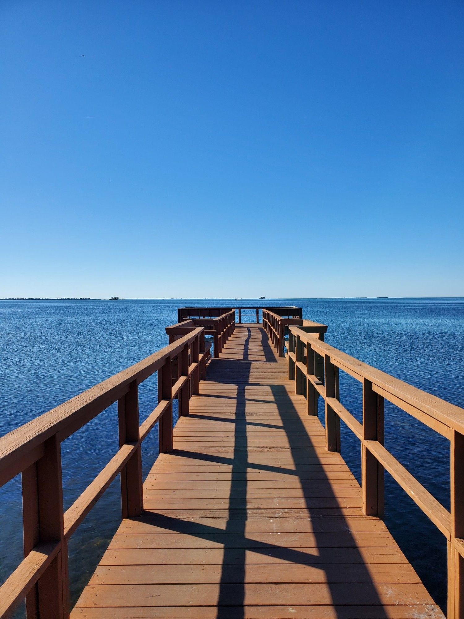 A stock photo of a wooden pier that stretches out into the calm dark blue waters. The blue sky is completely clear.