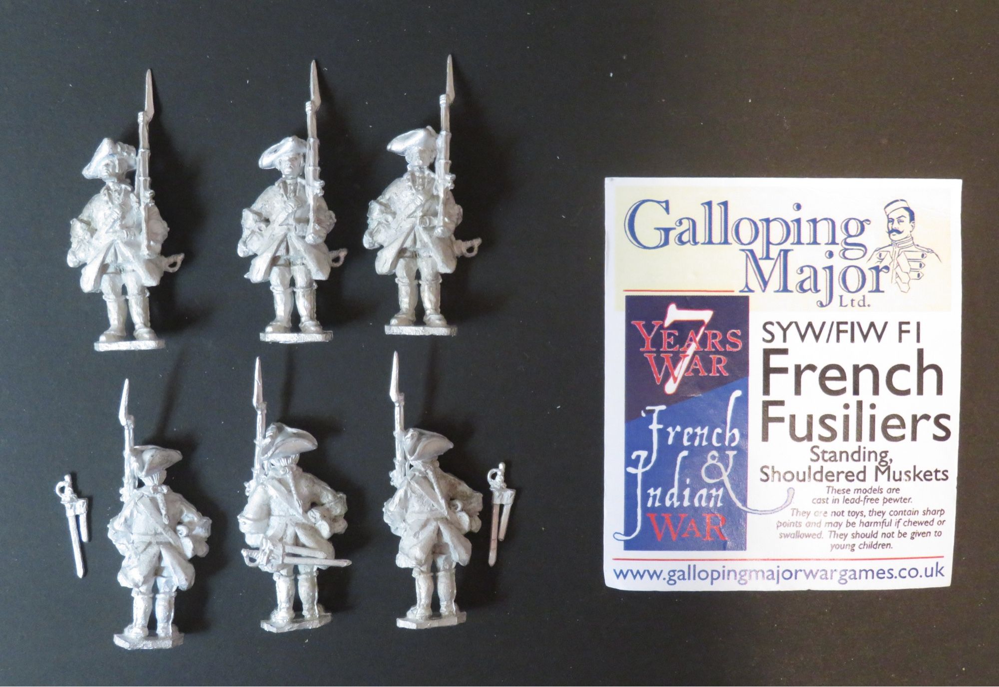 Photo of 28mm unpainted metal miniatures. 6 figures to left of photo in 2 rows of 3. They wear mid 18th century French military uniforms with tricorne hats and shouldered muskets. To the right of the photo is a header card with the manufacturer’s name (Galloping Major), brief description of the figures and contact details. All photographed on a black background.