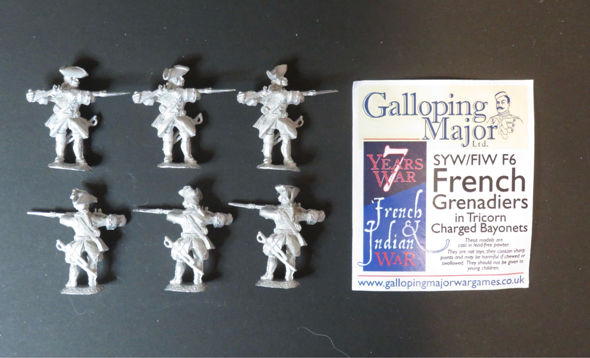 Photo of 28mm unpainted metal miniatures. 6 figures to left of photo in 2 rows of 3. They wear mid 18th century French military uniforms (Grenadiers) with tricorne hats and charged muskets with fixed bayonets. To the right of the photo is a header card with the manufacturer’s name (Galloping Major), brief description of the figures and contact details. All photographed on a black background.