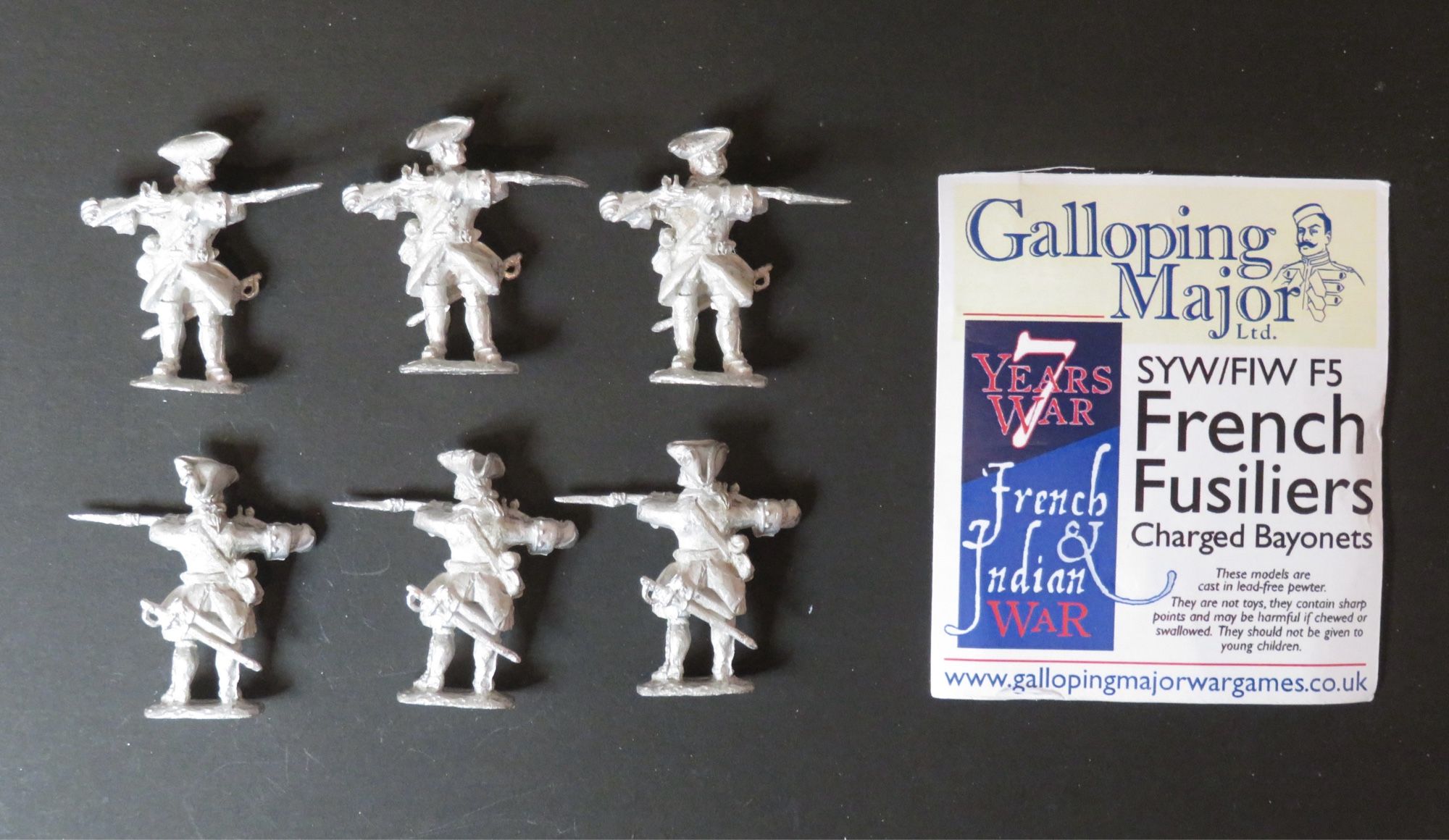 Photo of 28mm unpainted metal miniatures. 6 figures to left of photo in 2 rows of 3. They wear mid 18th century French military uniforms (Fusiliers ) with tricorne hats and charged muskets with fixed bayonets. To the right of the photo is a header card with the manufacturer’s name (Galloping Major), brief description of the figures and contact details. All photographed on a black background.