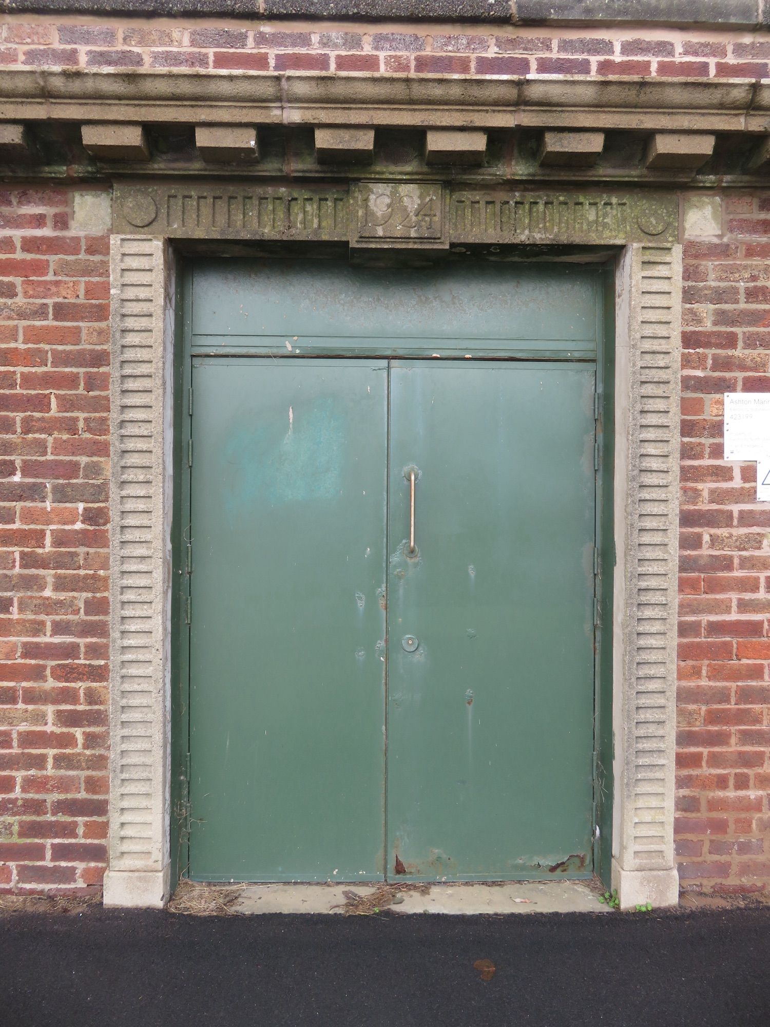 Photograph of a pair of green painted metal doors, set in metal sub frame with full width opaque over light also painted green. The sun frame is located within a brick wall and further framed by decorative precast concrete reveals. Above the doors and running horizontally across the full frame are further precast concrete sections forming a cornice with dentils. 

Location: Electricity sub-station, Fairhaven Lake, Latham St Annes, UK