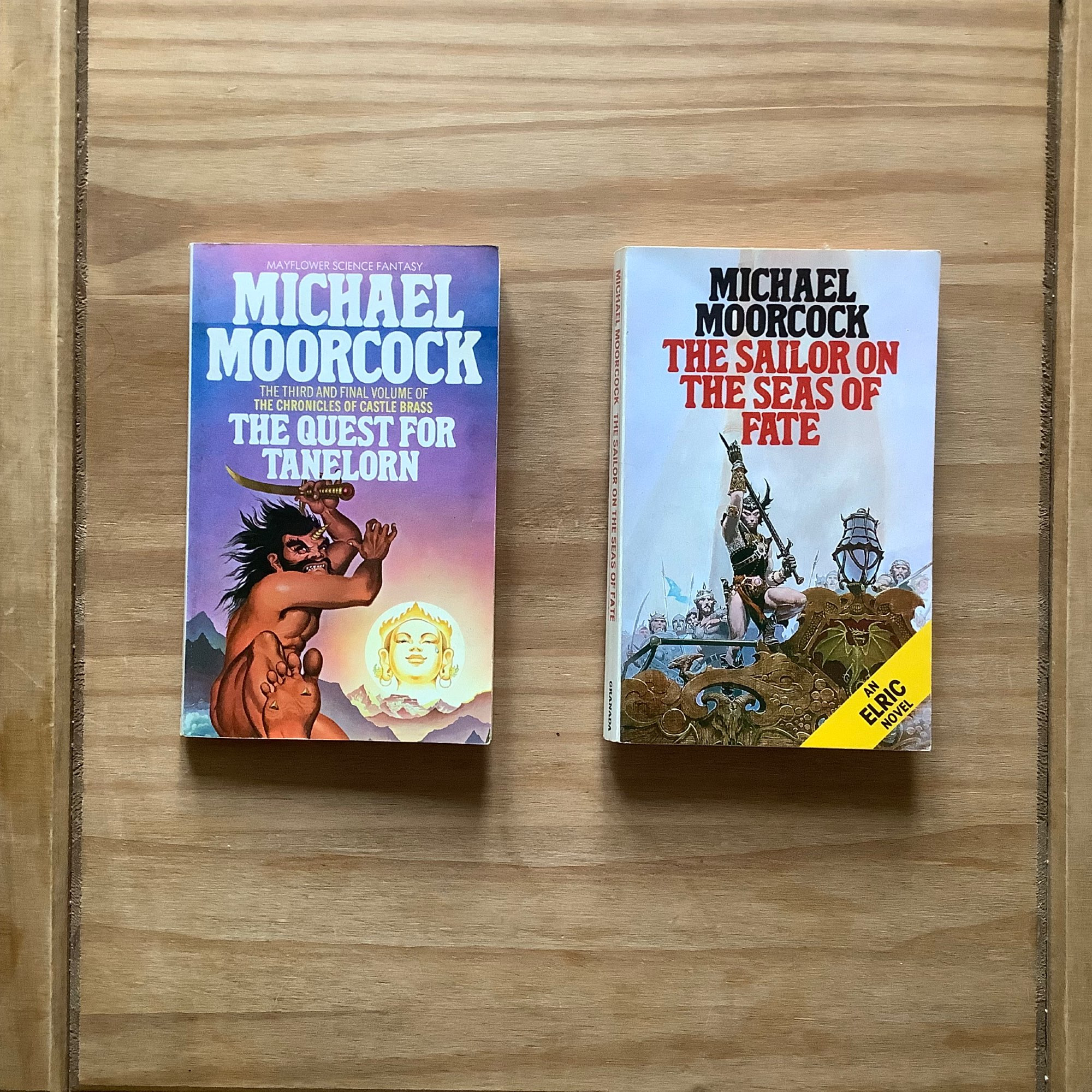 Photo of two classic Michael Moorcock paperback novels - “The Quest for Tanerlorn” with a fantastical cover art by Bob Haberfield. Also,the Elric novel “The Sailor on the Sea of Fate” with cover art by Michael Whelan.