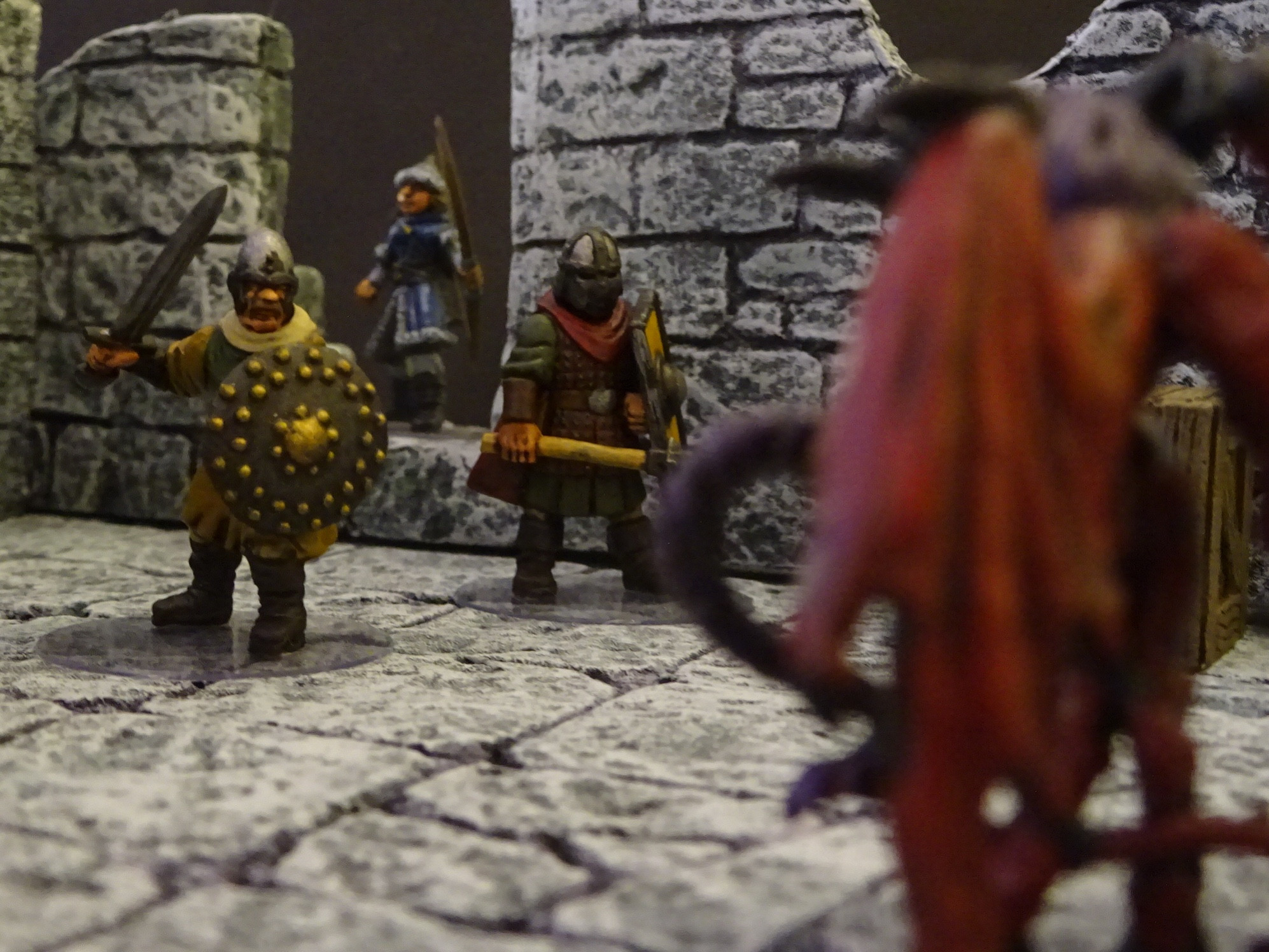 Photo of three fantasy adventures, facing towards the viewer, being confronted b a large , red demon, positioned to the right of frame. 

28mm multipart plastic Frostgrave miniatures, wearing pseudo Western Europe’s medieval clothing, and carry typical fantasy weaponry. They are in a ruined environment.