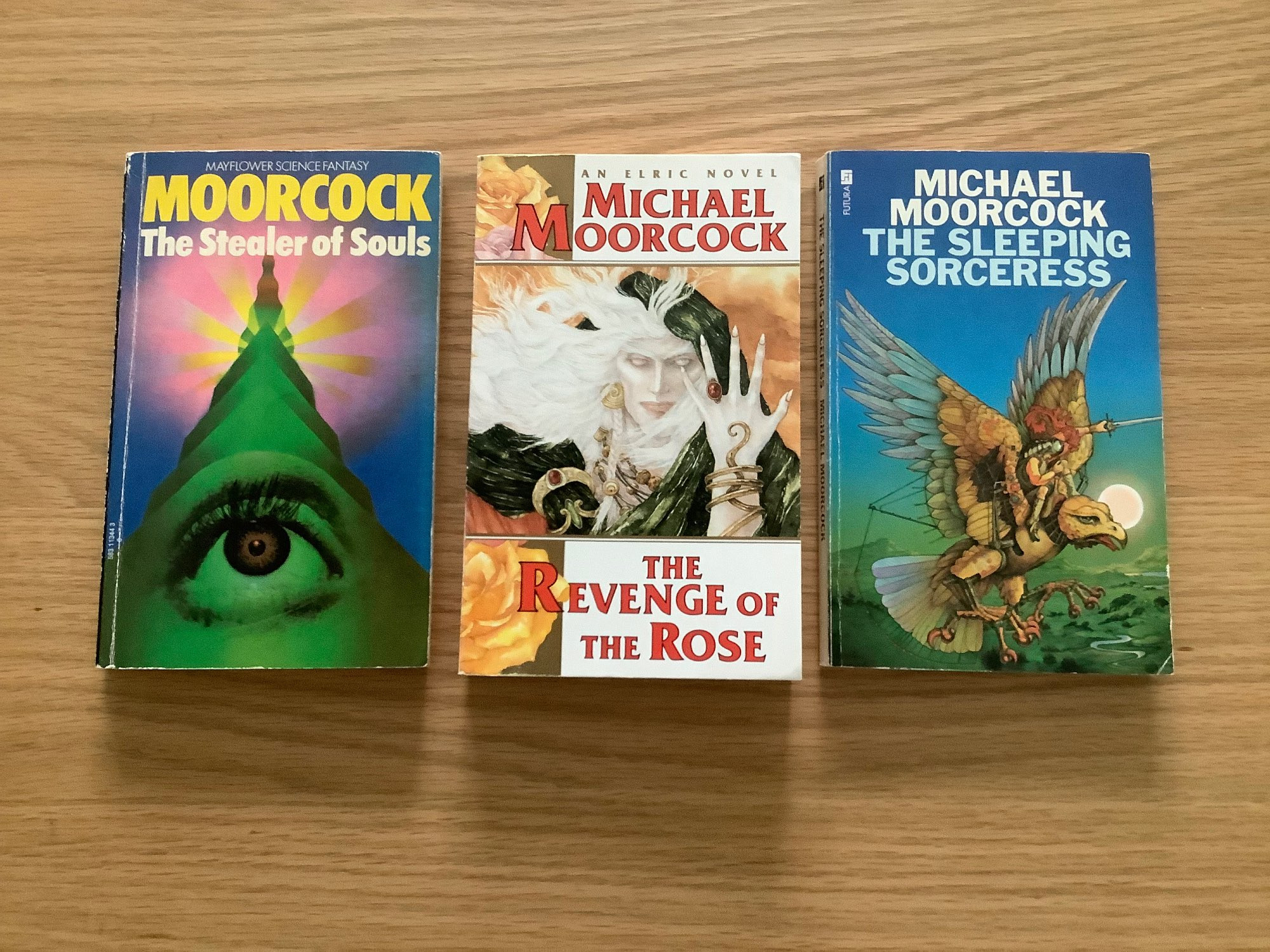 Three paperback books by Michael Moorcock. 

On the left is “The Stealer of Souls” with cover art by Bob Haberfield. It depicts a green tower (?) as viewed from the ground, against a pink fading to blue sky. Towards the top of the tower are rays of light. An eye looking upwards is incorporated into the lower part of the structure. 

The centre book is “The Revenge of the Rose” with cover art by Robert Gould. It depicts Elric as a white haired, pale skinned being with his left hand raised level with his face, fingers splayed. His long hair streams to the left of the cover. 

The right hand book is “The Sleeping Sorceress” with cover art by Patrick Woodroffe. It depicts a bird like creature, possibly part robot or cyborg, with wings up stretched against a green base fading to blue background. A knight like figure sits astride the creatures neck.