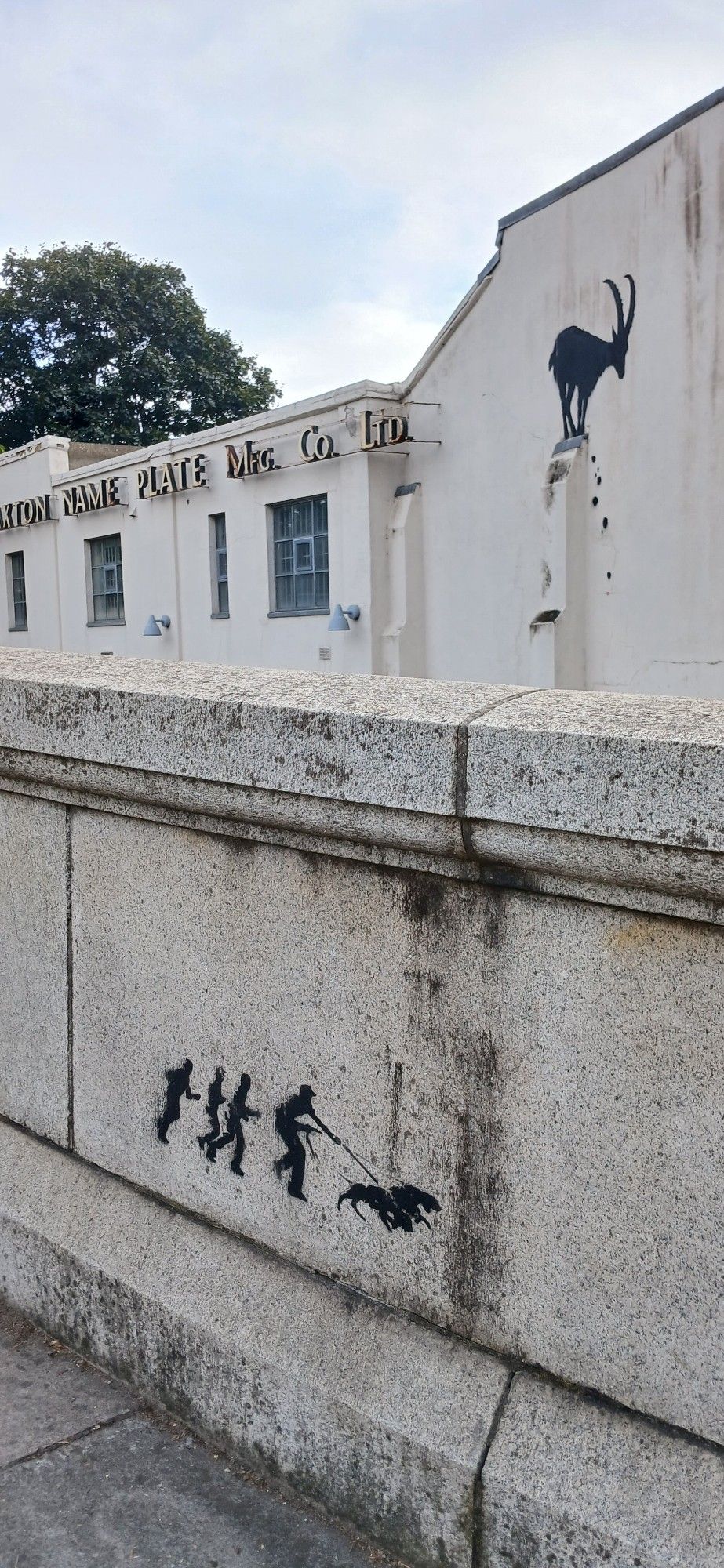 Banksy art work on white walls.  Black silhouette of a goat balancing on top of a beam. Some black rocks appear to be falling.  In the foreground on a marble bridge railing a small artwork if a man with two dogs straining on their leads, followed by 3 other men.