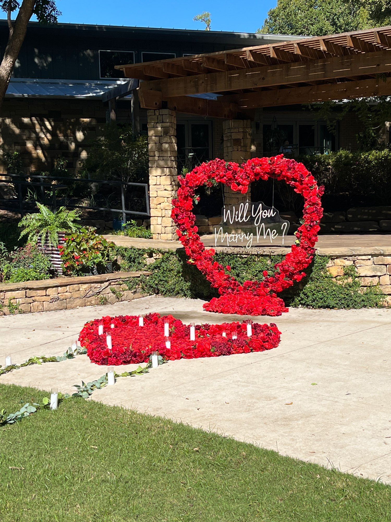 a giant heart made of red flowers in front of a small stone stage in a garden. there’s another large heart of flowers on the ground with candles around the edges. inside the standing heart are suspended the word “will you marry me”