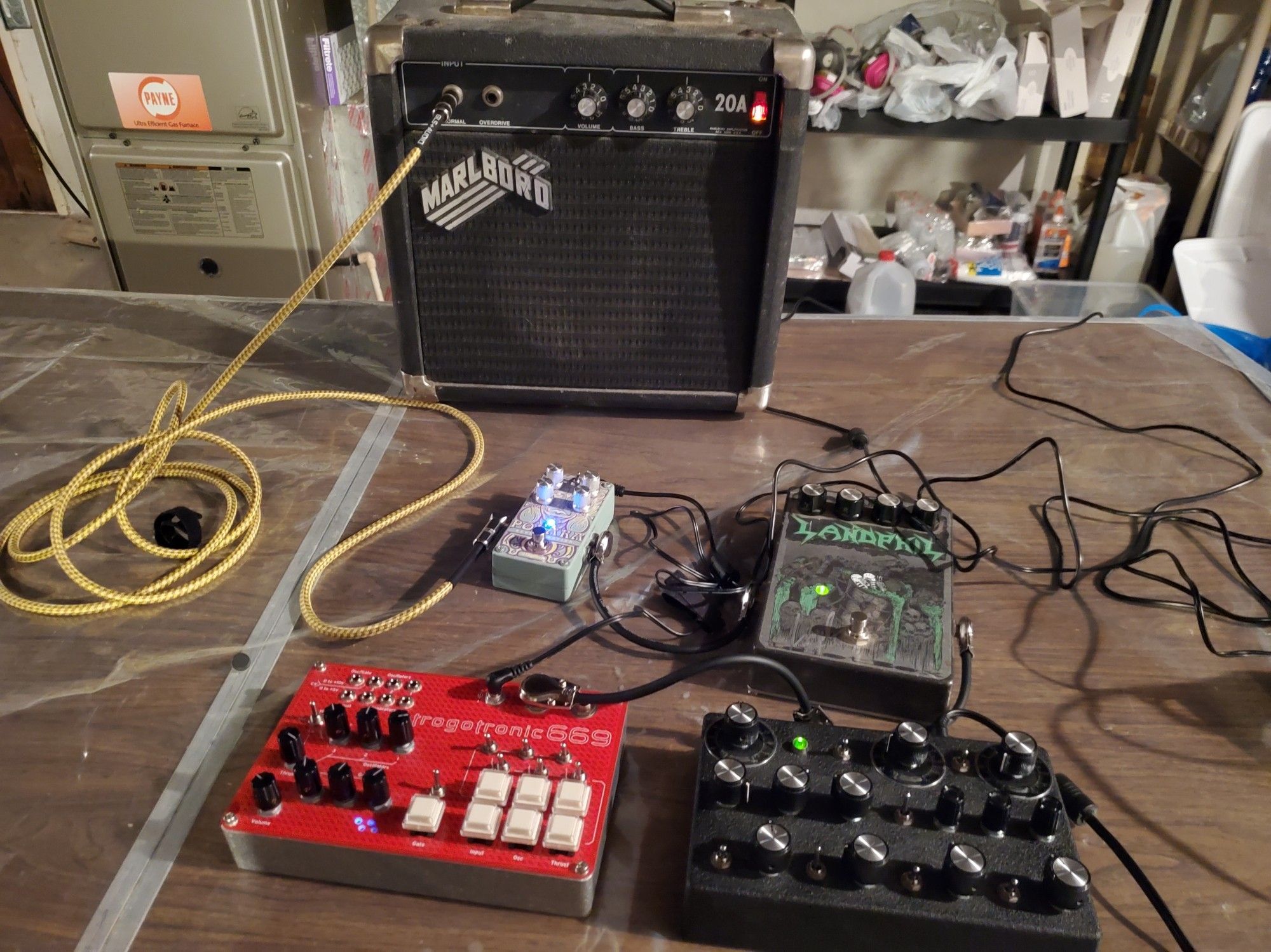 A Trogotronic 669cv Boss Hog MiniSynth chained into a JMT Synth DNVO-1 Desktop Synthesizer chained into an Idiotbox Landphil pedal and a Digitech Polara reverb, all plugged into a tiny, ancient Marlboro cigarettes branded practice amp.