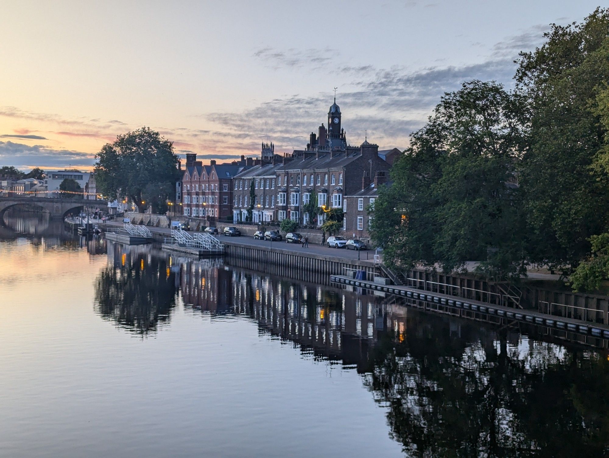 A picture of old buildings reflecting in the river Ouse. Pastel sunset, soft shadows, and calm water. Many ancient oaks frame the buildings