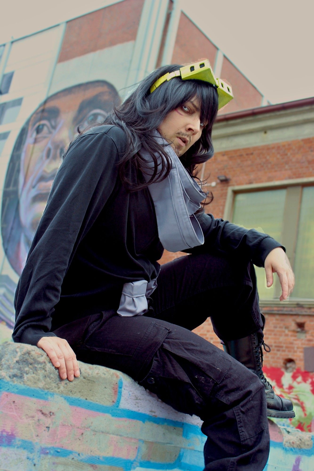 Me in a Aizawa Shota cosplay. I'm wearing black clothes and has yellow glasses on my head. I'm sitting on a brick wall in front of graffities and looking at the camera.