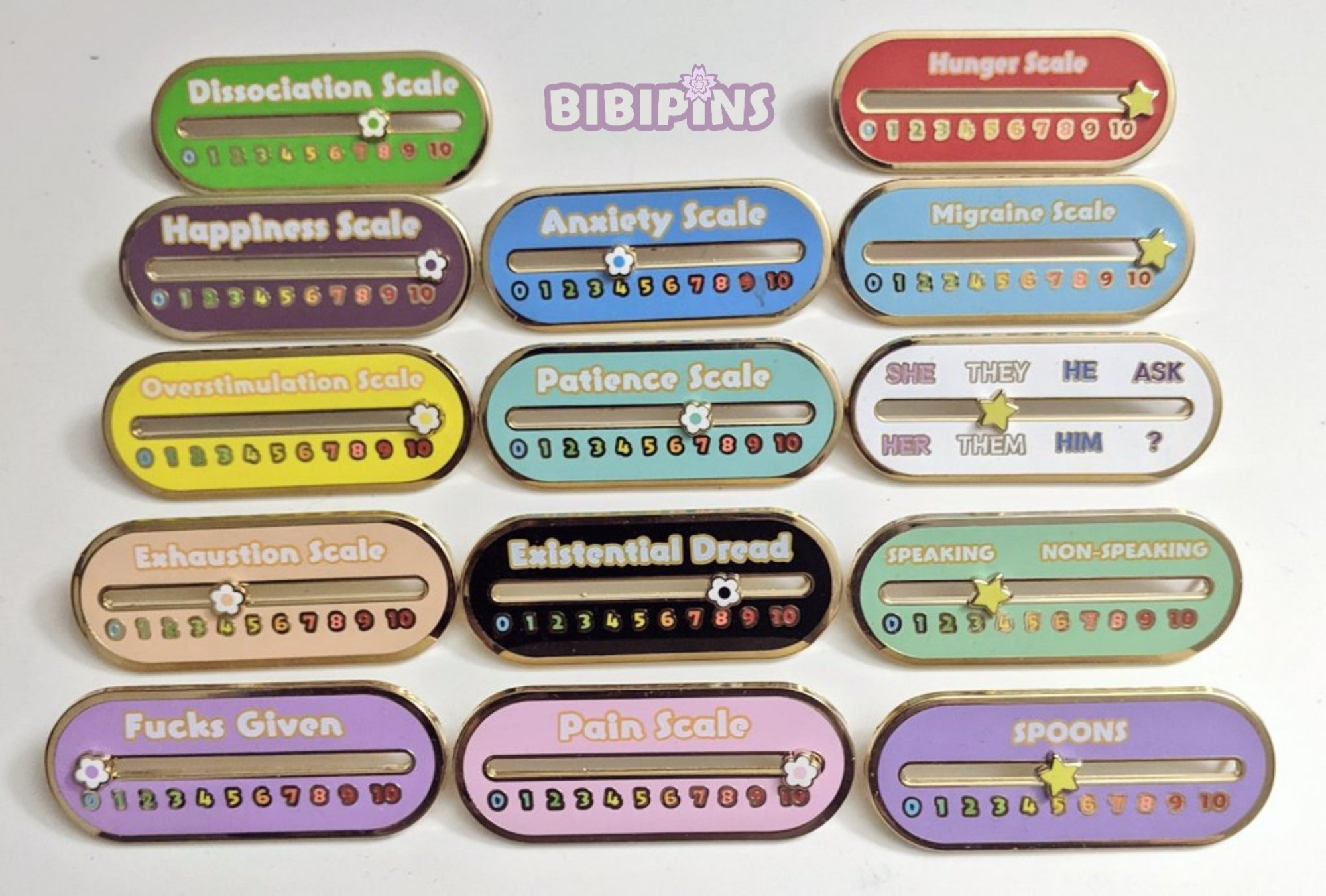 5 Oval shaped pins with gold outlines with an oval cutout in the middle with a yellow slideable star:

Spoons: Lavender background
Hunger Scale: Red background
Migraine Scale: Blue background
Speaking vs Non Speaking: Green background

The first one has SHE, THEY, HE on top. At the bottom there's HER, THEM, HIM. White background.