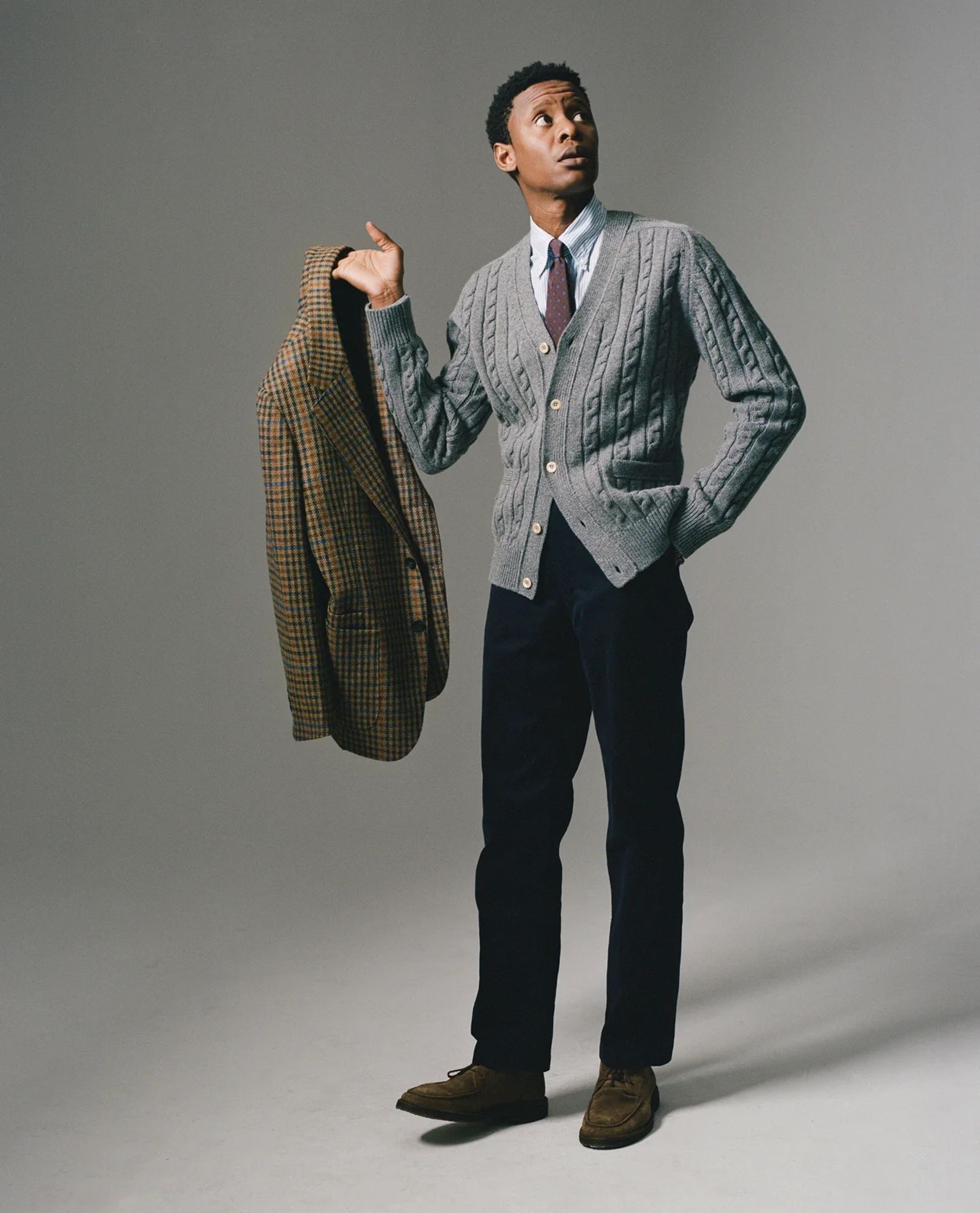 Black Drake's model looking dapper in navy trousers, a cable knit grey cardigan, a blue OCBD, a maroon tie, and holding a houndstooth sport coat