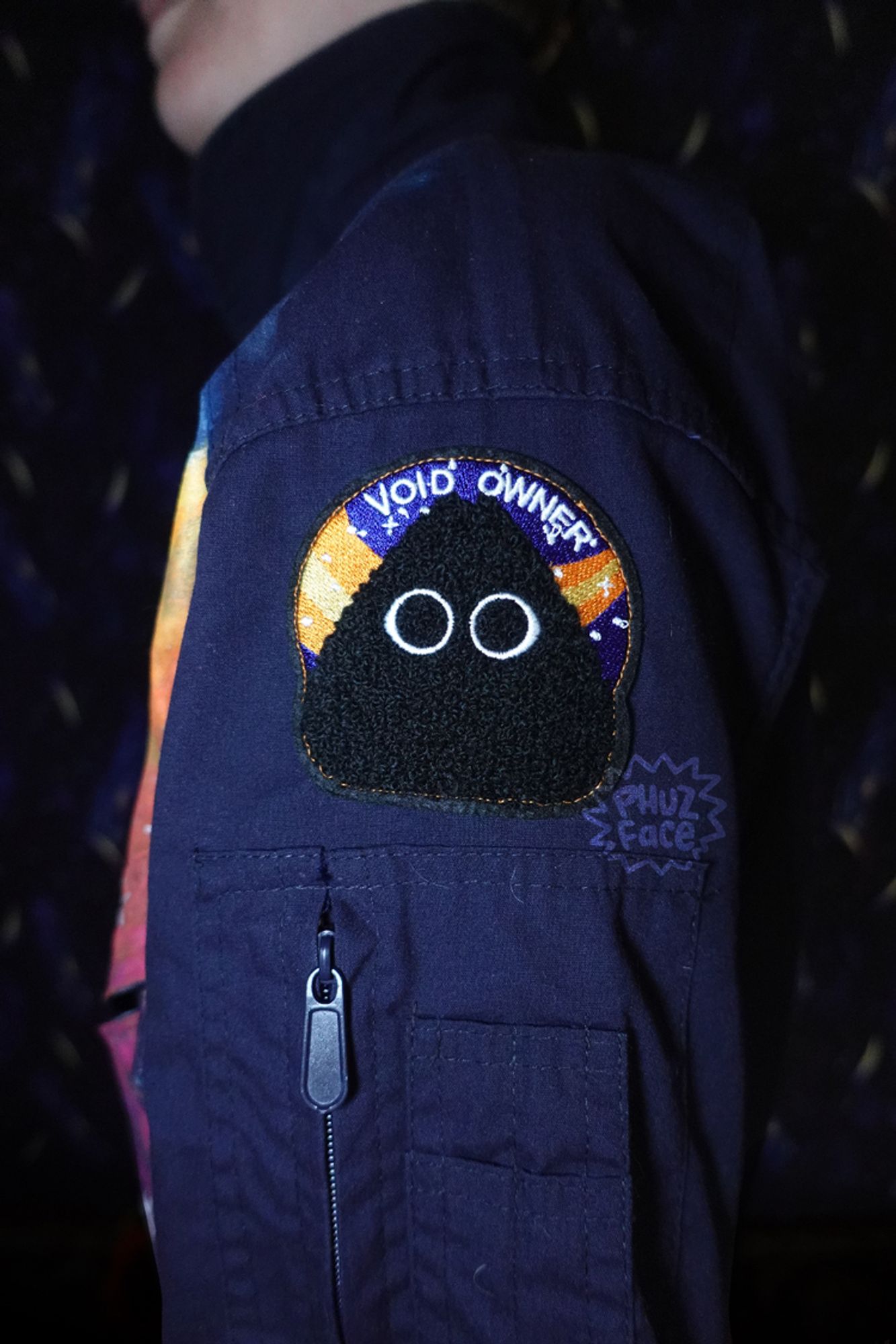 A Void Owner patch displayed on a navy blue flightsuit's shoulder, being worn by the artist. The dark purple blue cosmic tablecloth is being used as a backdrop.