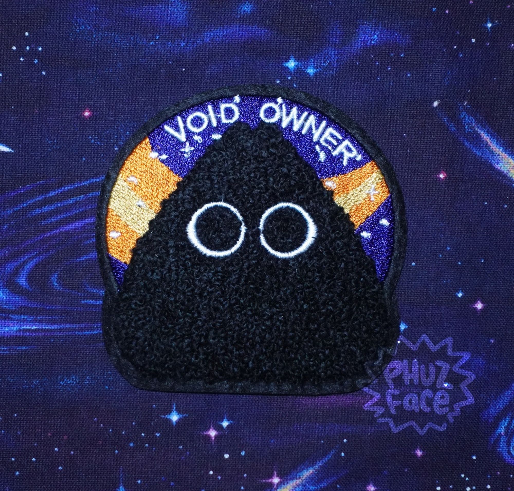 Picture of a space mission badge-styled iron-on patch featuring a stylized chenille black cat in front of embroidered dark blue circle with a yellow and orange comet swoosh and small white stars. White text above cat reads "VOID OWNER." The patch is on a dark blue and purple cosmic tablecloth with stars, galaxies, and comets.