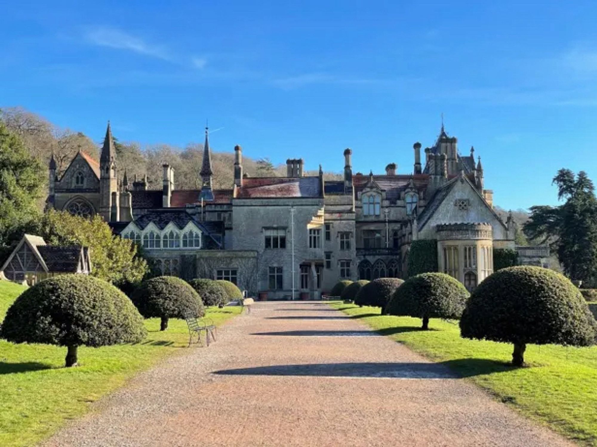 A path running towards an ornate stately home, with lawn and topiary either side of the path, and blue sky above.