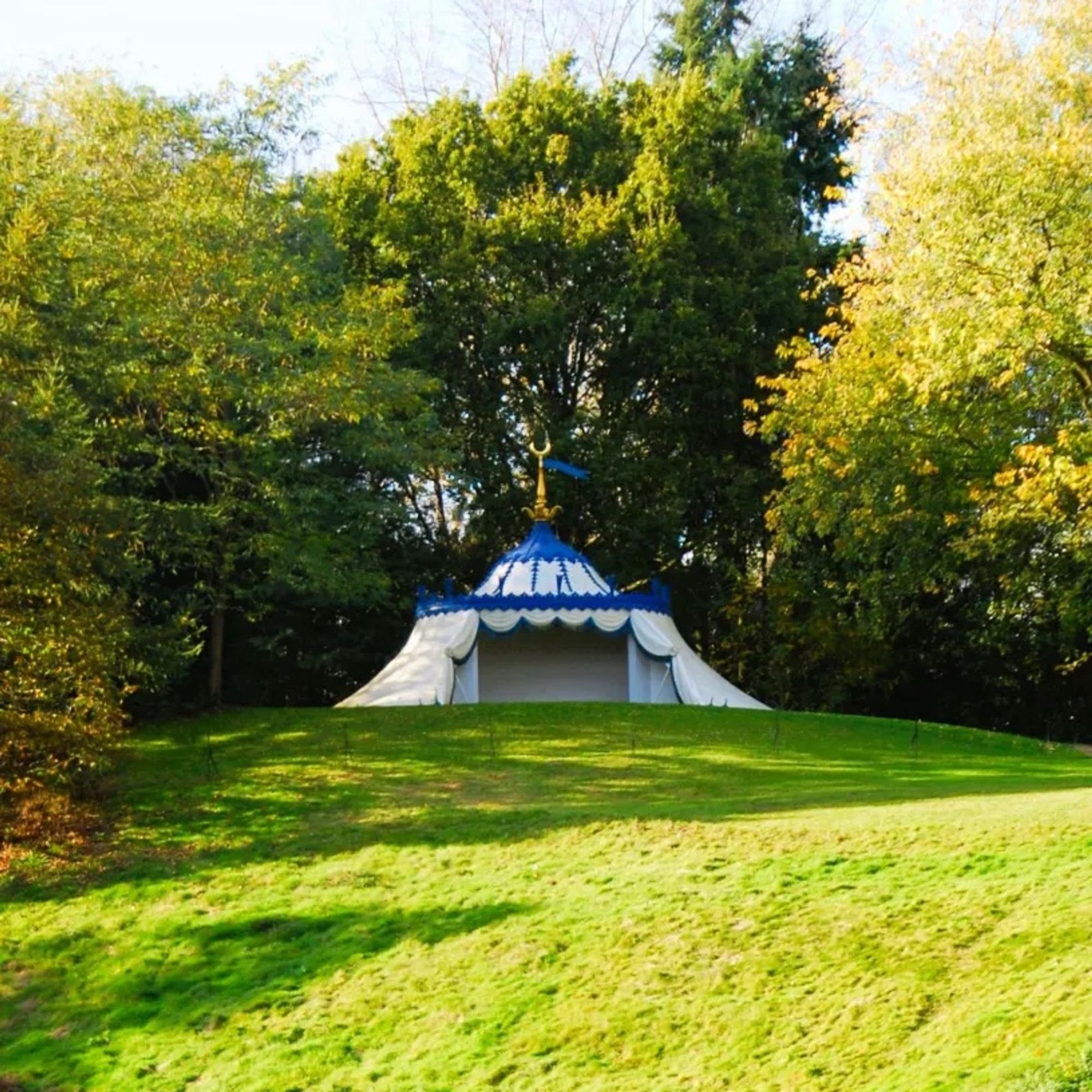 An ornate white tent with blue trim, at the top of a hill, with trees behind it.