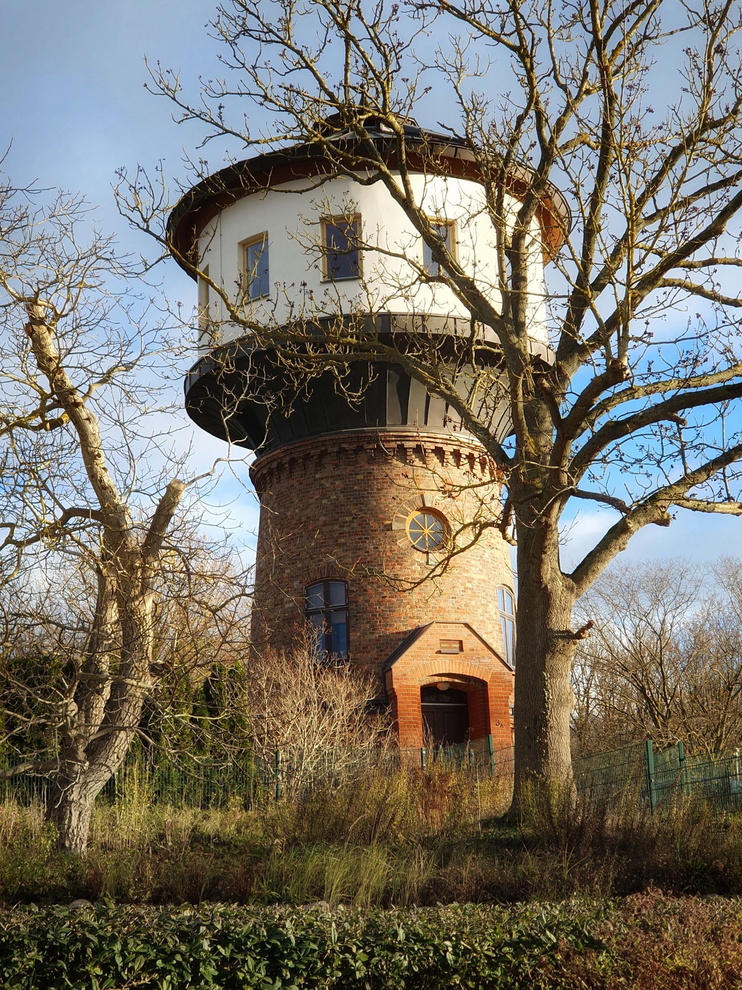 Ein einzelnes rundes hohes Gebäude in Form eines Leuchtturm aus rotem Backsteinen. Der obere Teil ist weiß mit viel Fenstern. Vr und hinter dem Turm sind blätterlose Bäume zu sehen. Im Vordergrund eine grüne Bepflanzung. Der Himmel ist blau mir Schleierwolken.