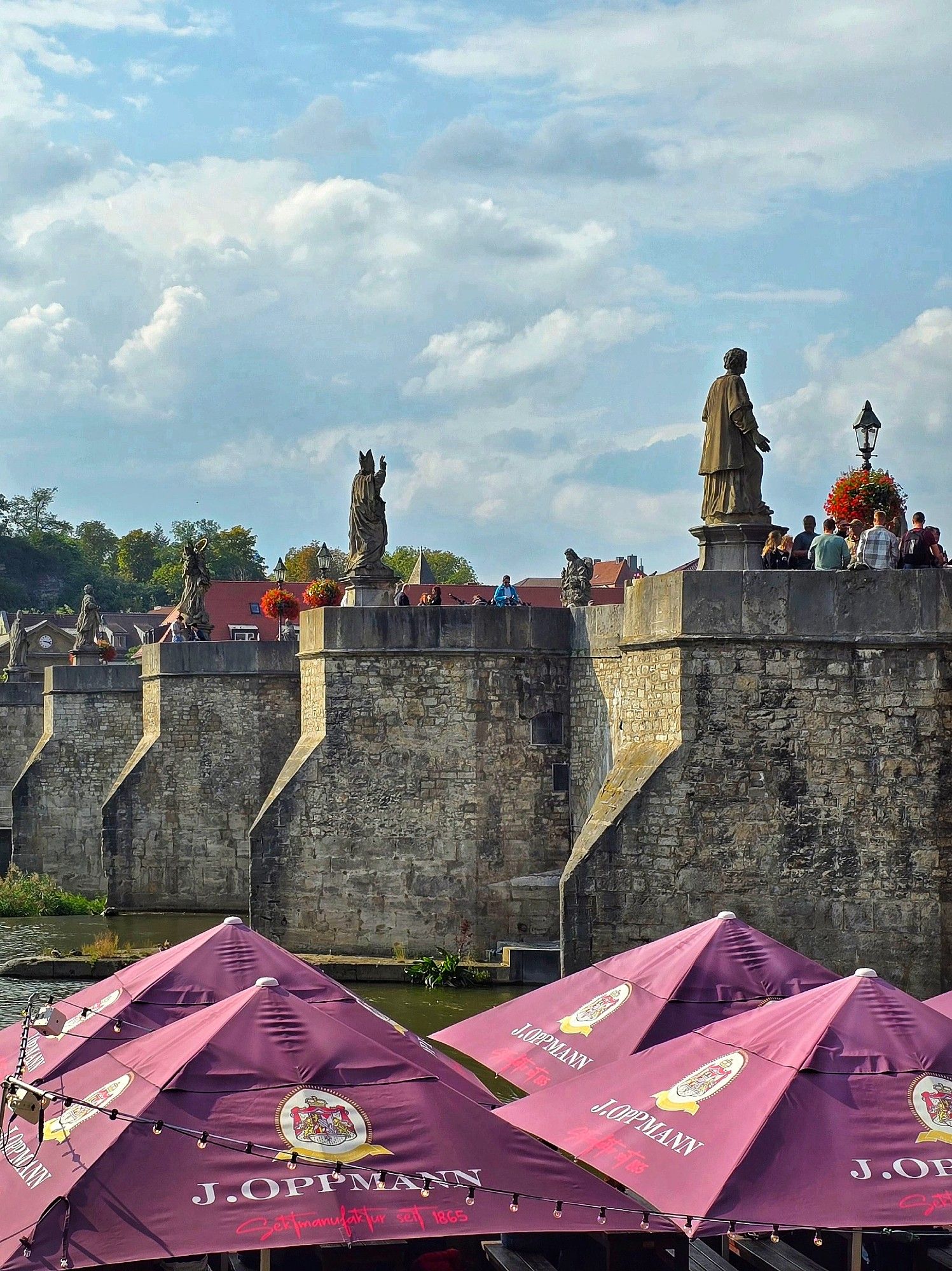 Eine alte Brücke mit Sulpturen aud der viele Menschen stehen. Im unteren Teil des Bildes sind violette Sonnenschirme mit Werbeaufschrift zu sehen. Der Himmel ist blau mit vielen großen Wolken. Zwischen den Schirmen und der Brücke ist ein bisschen von einem Fluss zu sehen.