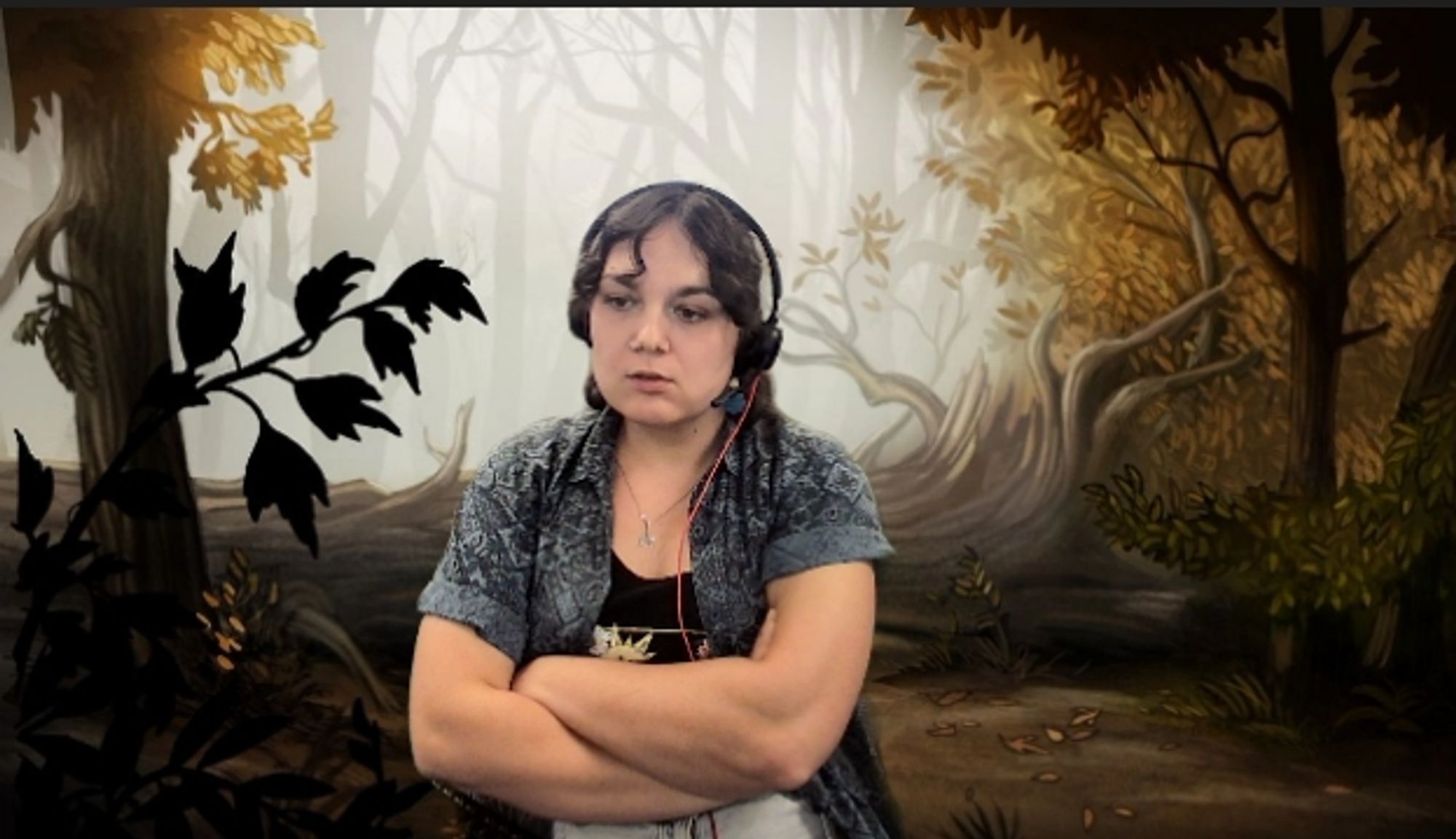 a woman on a video call with an autumnal background. her arms are crossed, revealing her muscles, she is wearing a blue overshirt with a black tank top