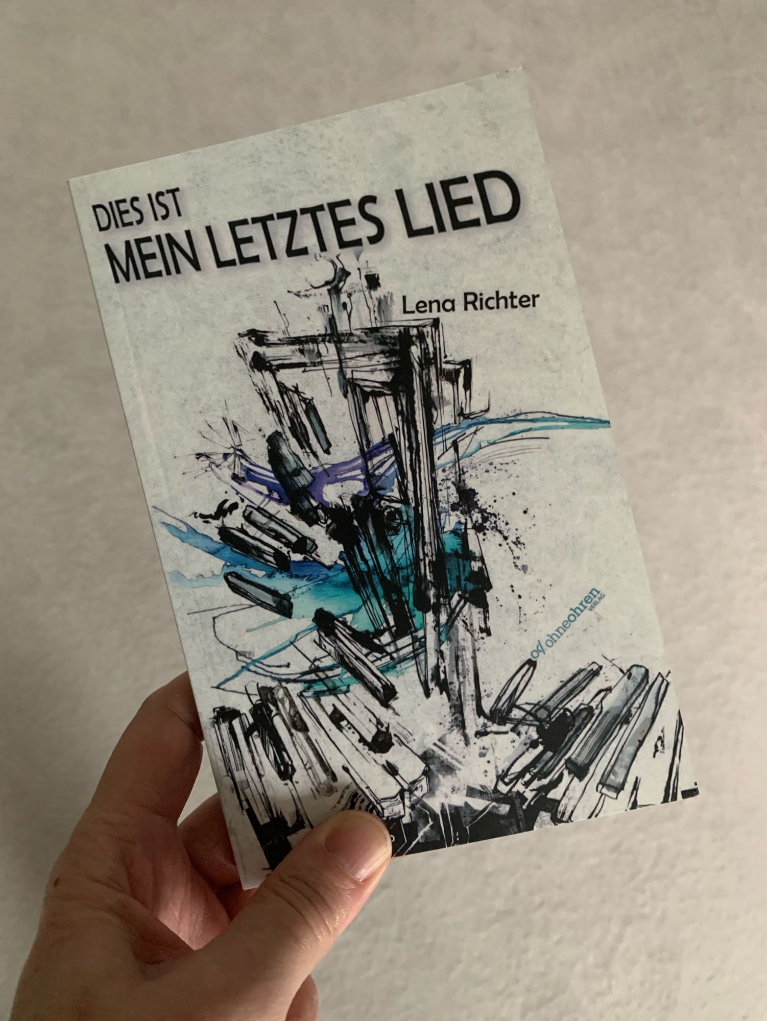 Taschenbuch “Das ist mein letztes Lied” von Lena Richter aus dem ohneohren-Verlag. Auf dem weißen Cover ist eine abstrakte Zeichnung in Schwarz und Blau, die mich an einen auseinander fallenden Turm aus Stelen erinnert.