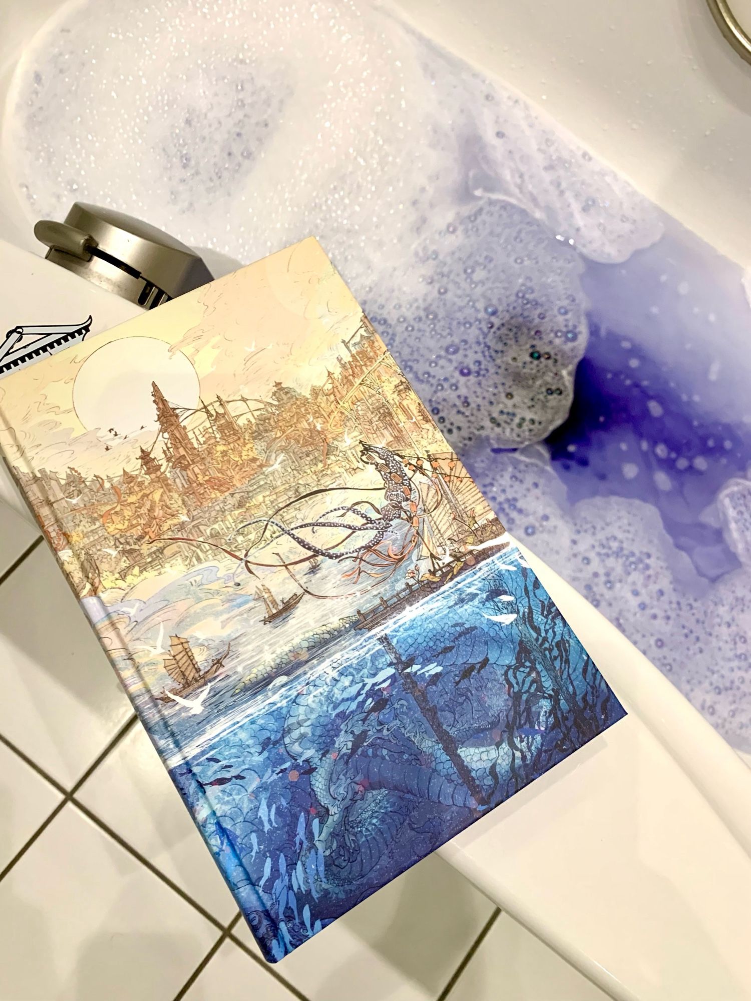 Eine Badewanne mit blauem Schaumbad. Auf dem Rand liegt eine Illumicrate Ausgabe von „Fathomfolk“ von Eliza Chan. Das naked hardcover ziert eine fein gezeichnete Fantasywelt mit einer südasiatisch anmutenden Stadt, die über einer bevölkerten Unterwasserwelt thront.