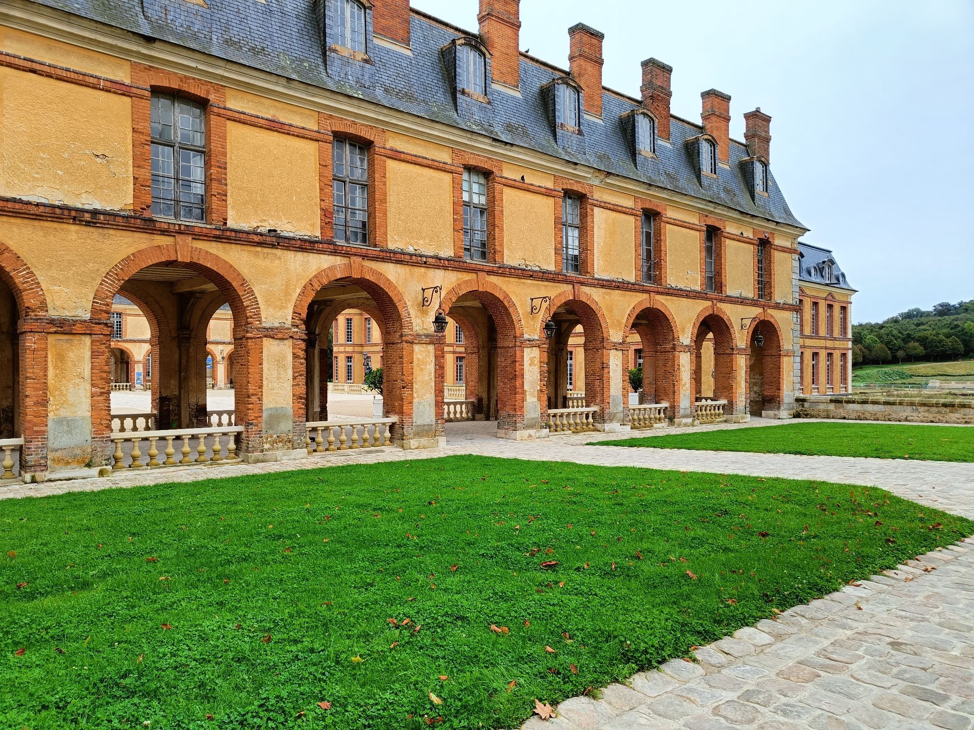 An orangeish building with arches on the ground floor and lawns in front with a light path leading to it and a courtyard behind