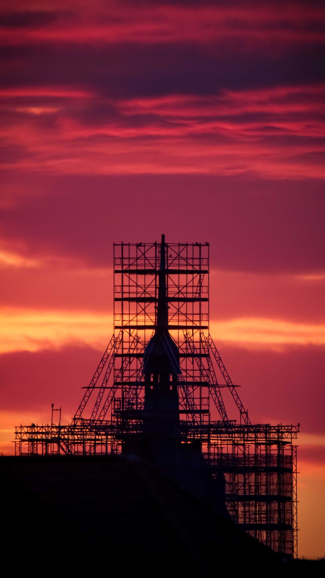 Rot, lila, Orange sind die Farben am Morgenhimmel. Zoom über die Dächer. Silhouette eines kleinen Turmes auf einem Dach, welcher komplett von der seinem Gerüst - Skelett umgeben ist. Foto vom Mann zur Verfügung gestellt.