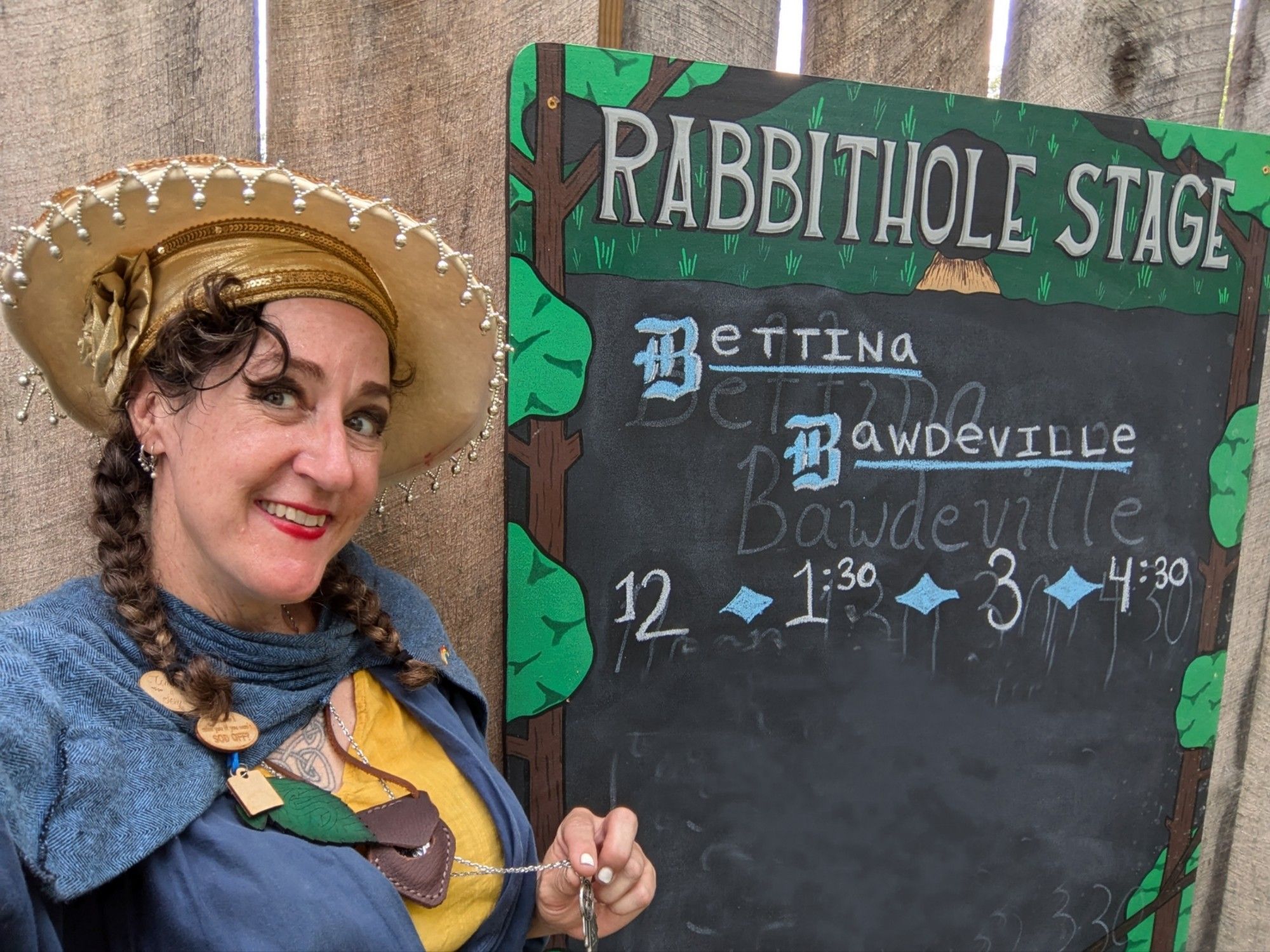 Bettina wears her signature fancy wide brimmed cream colored wool hat with shiny gold trim. She holds up a TARDIS key and looks thrilled to be standing next to the sign for the Rabbit Hole stage with her showtimes: Noon, 1:30, 3 and 4:30